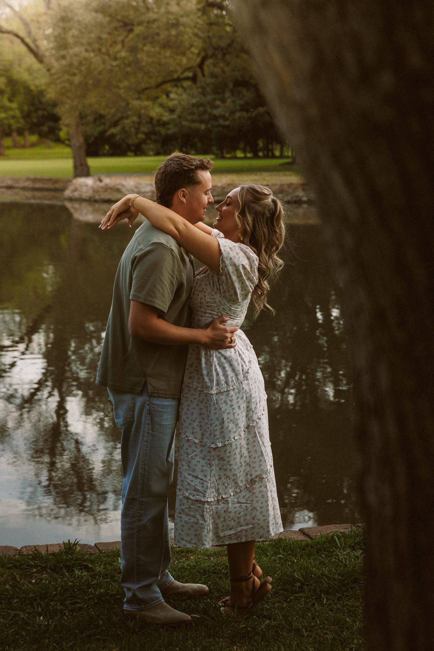 Couple embracing by a reflective pond in a lush green park during golden hour.