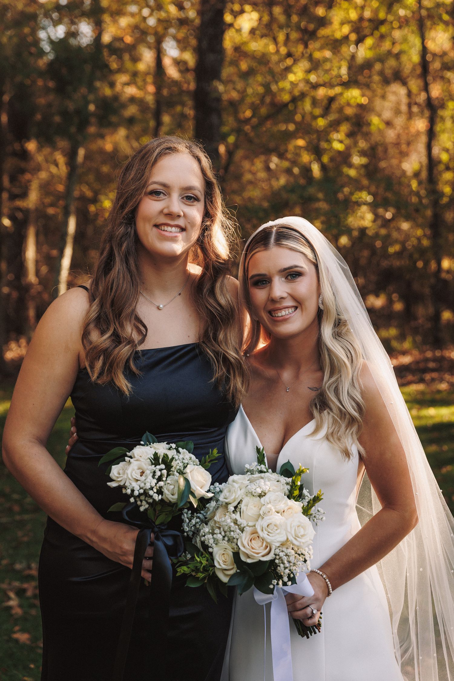 Bride in white gown and bridesmaid in black dress hold white rose bouquets in a sunlit autumn forest setting.