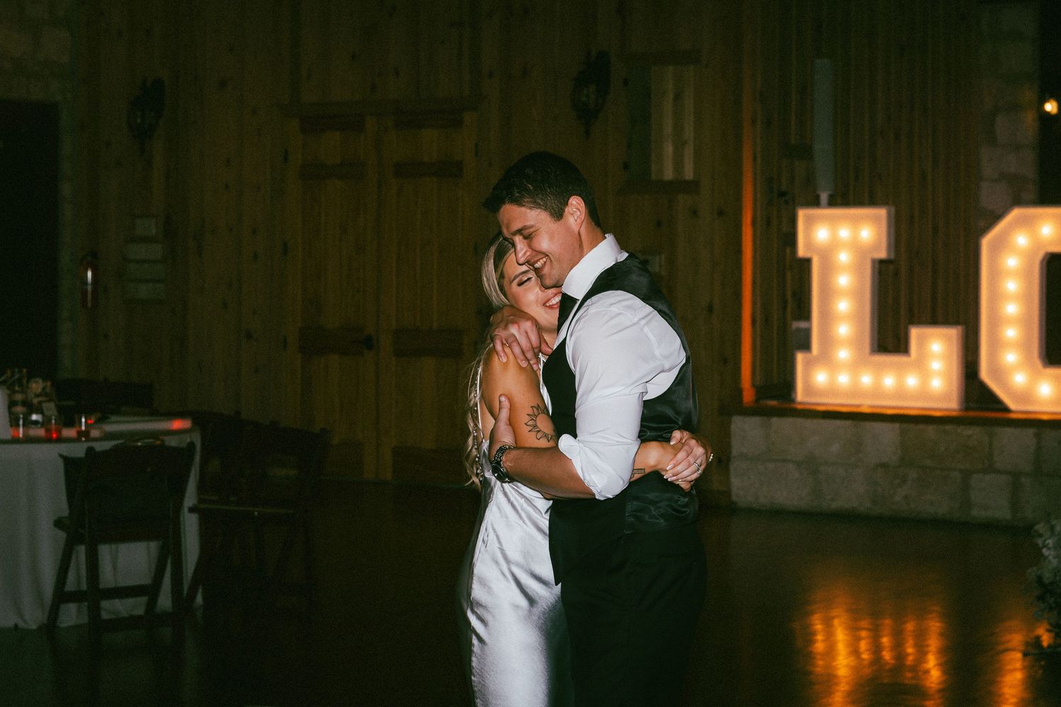 Bride and groom share romantic first dance in warmly lit rustic barn with marquee letter backdrop.