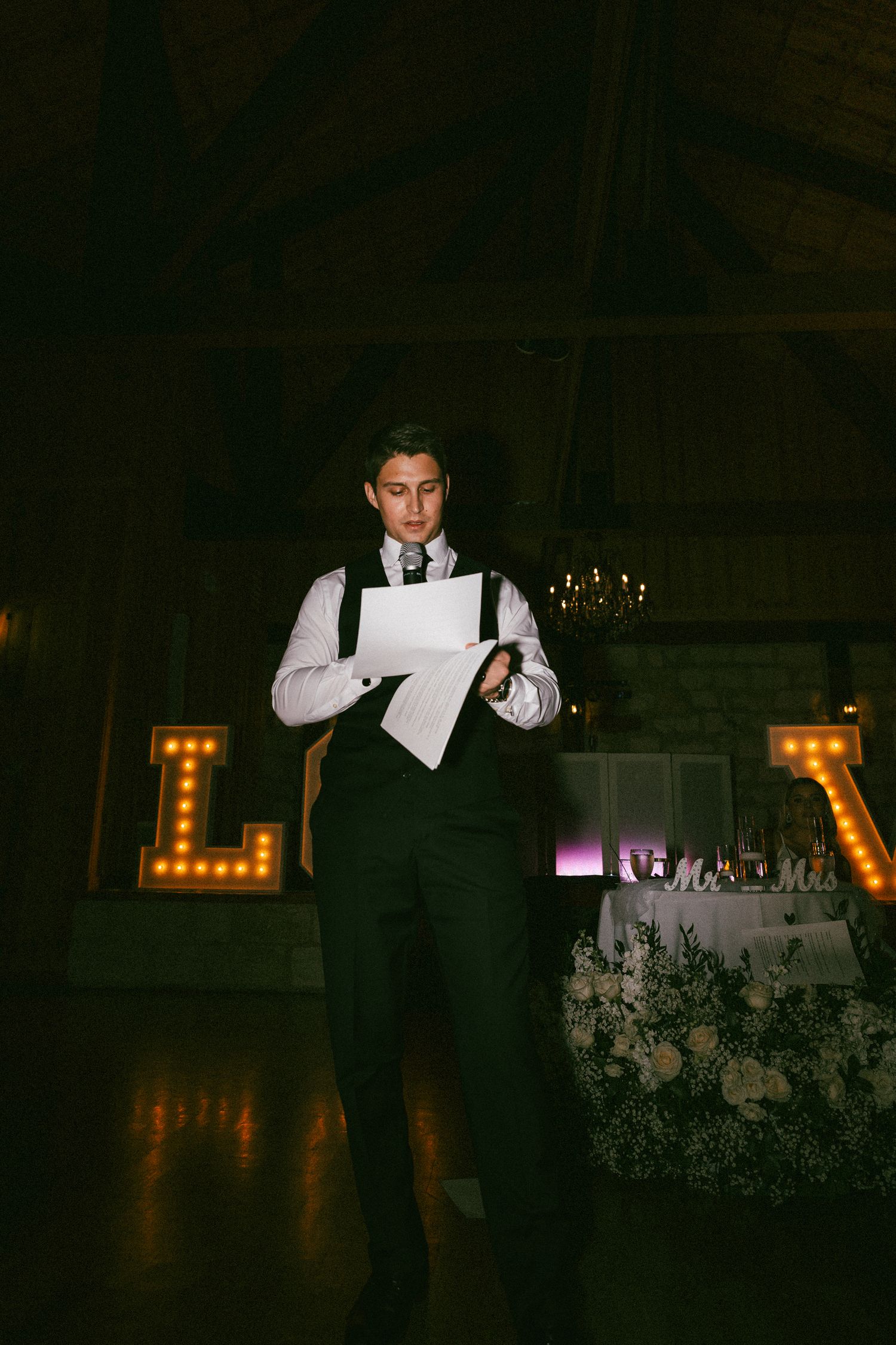 A young man in a vest reads a wedding speech from papers in a dimly lit reception hall.
