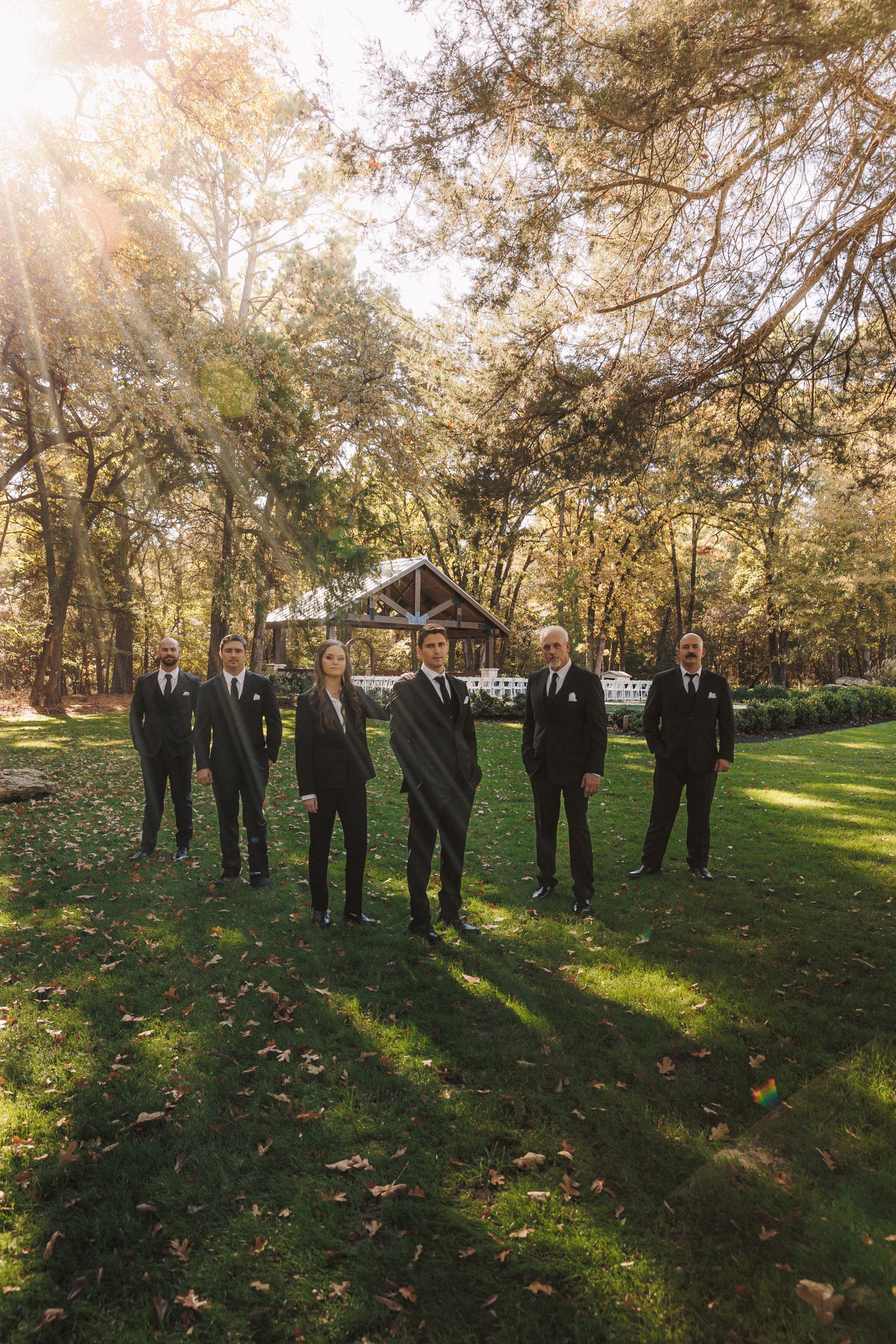 Groomsmen in black suits stand together on green grass at an outdoor woodland wedding venue on a sunny autumn day.
