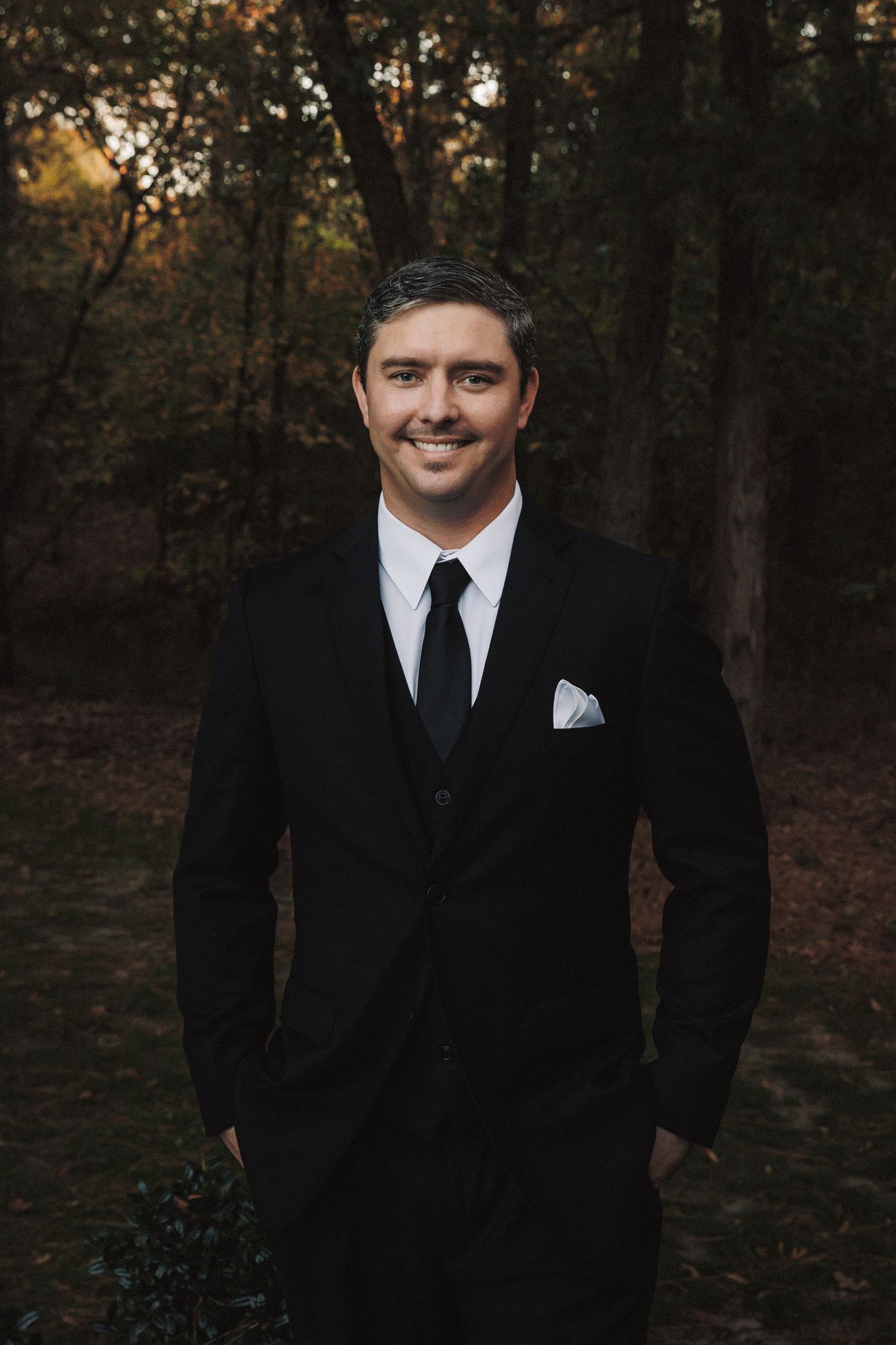 Handsome groom in black suit with tie and pocket square smiling in autumn forest setting.