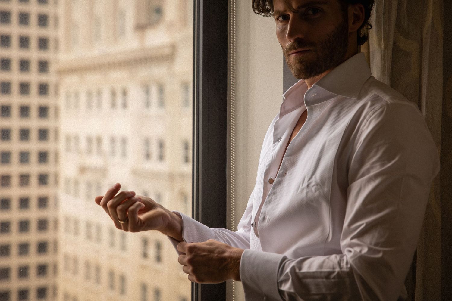 Groom in white dress shirt buttoning his cuff links while standing by a large city-view window.