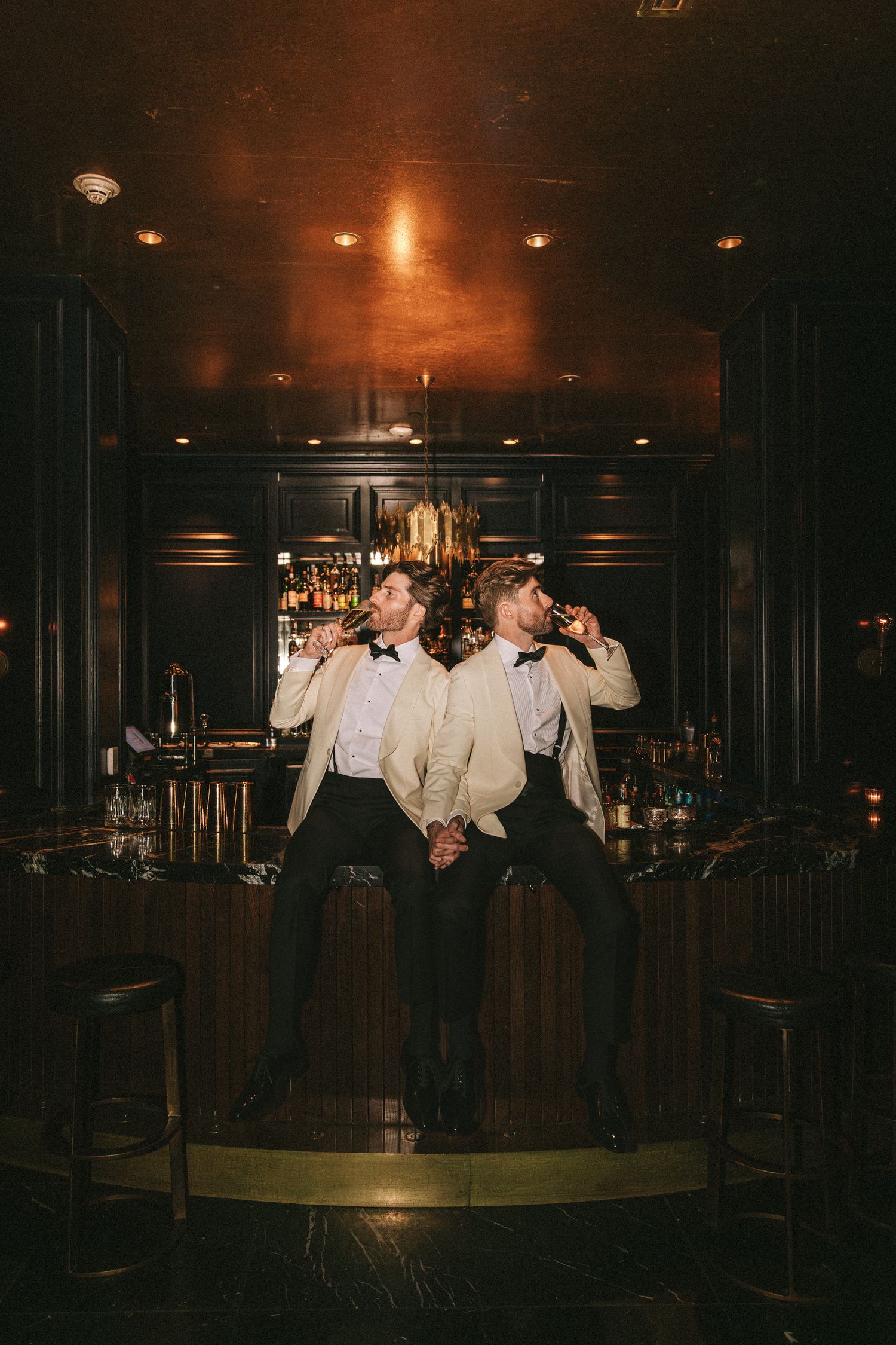 Two grooms in matching ivory tuxedos sitting at dark bar, drinking and holding hands at wedding.
