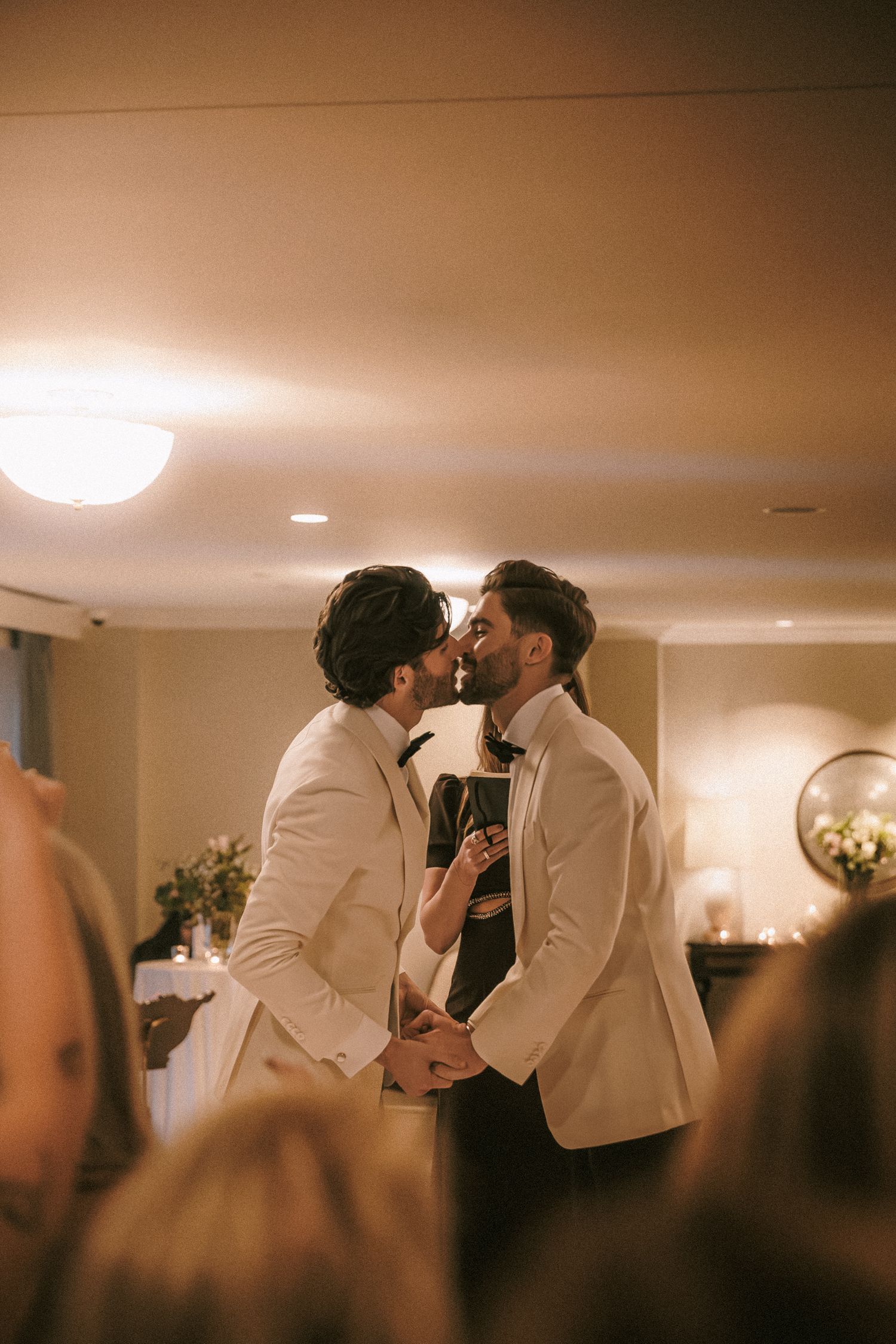 Two grooms in white tuxedos share a first kiss at their wedding reception.