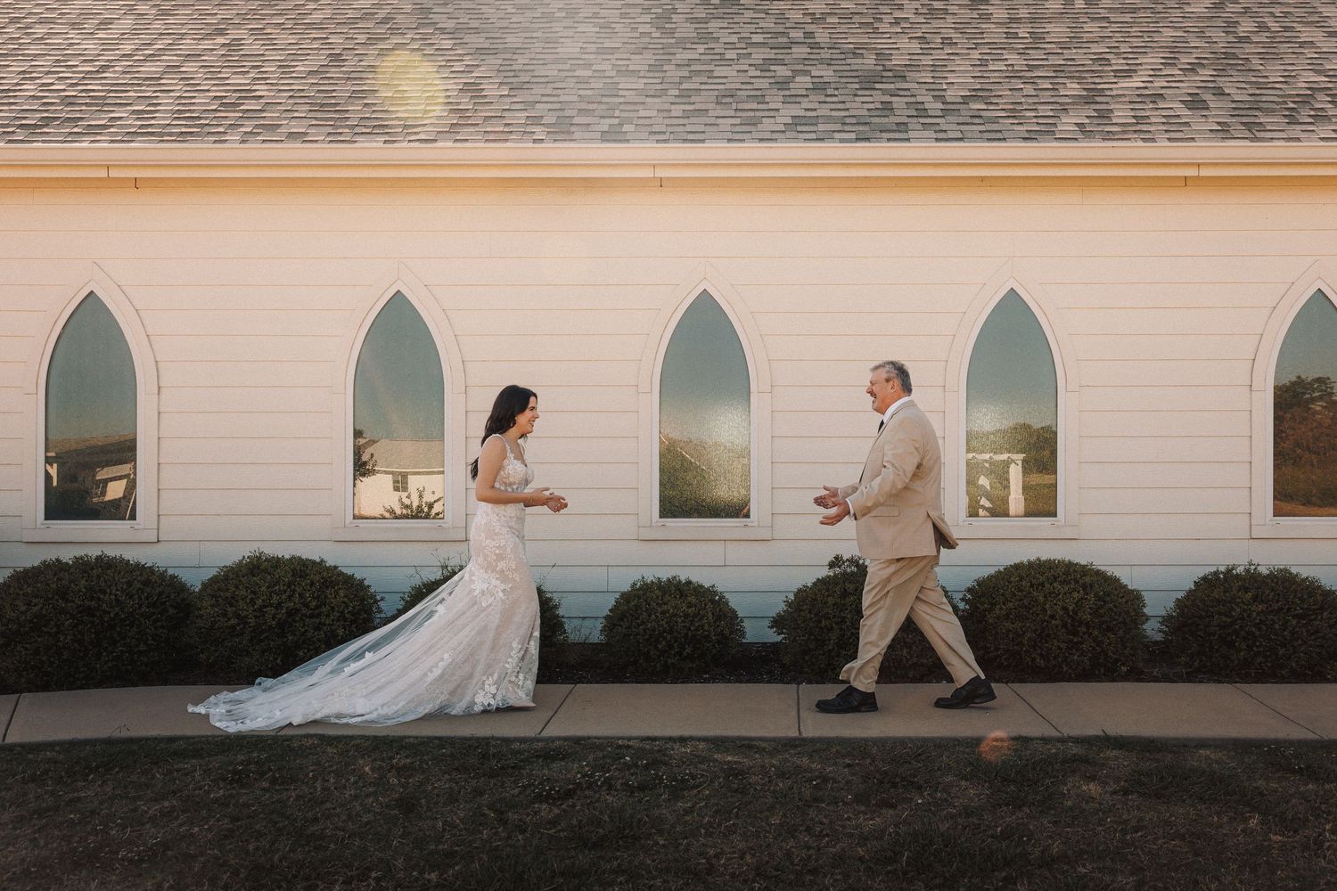 Bride in lace gown with train walks toward man in tan suit outside white chapel at golden hour.