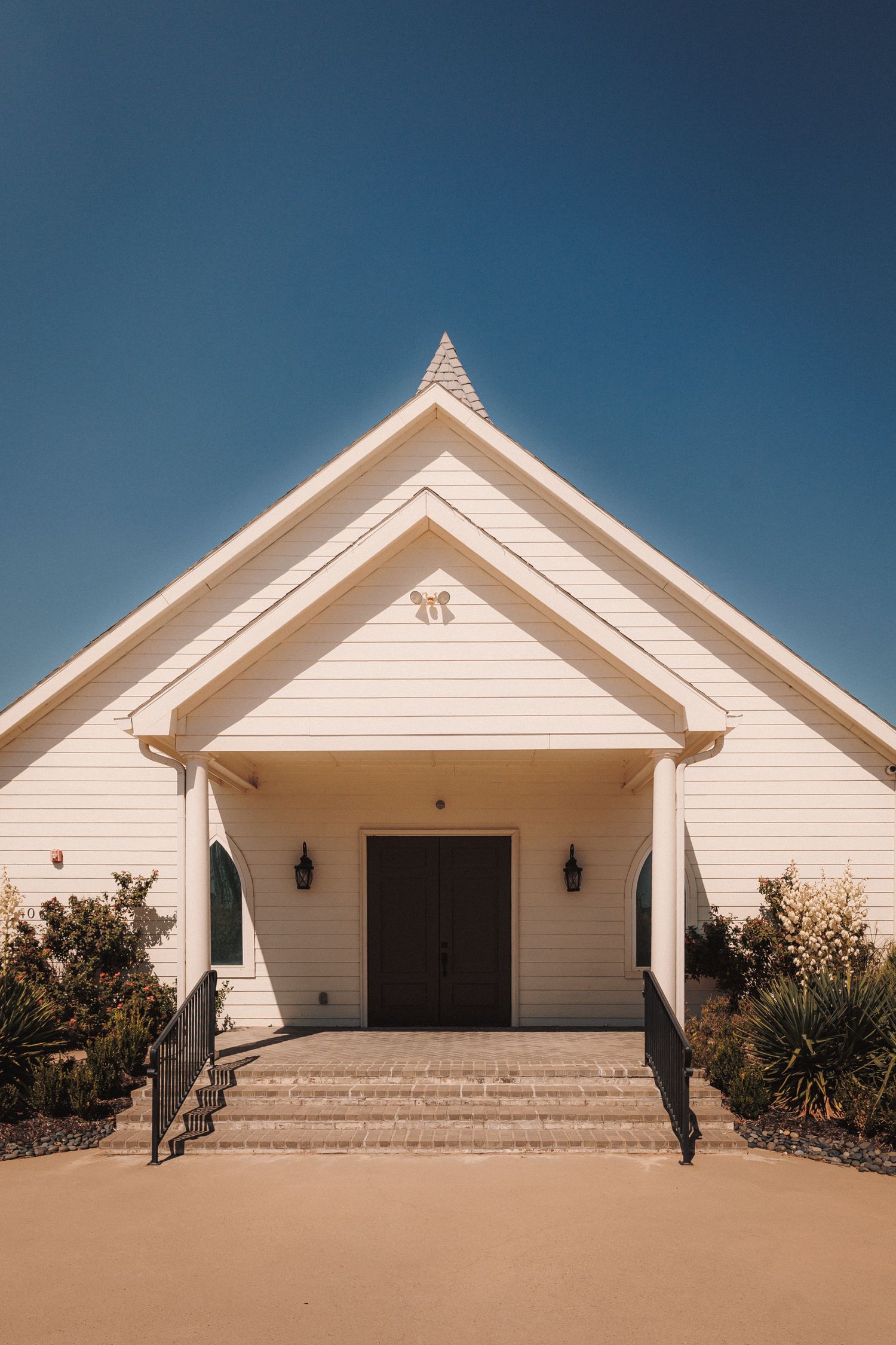 White wooden chapel with steeple, arched entrance, lantern sconces, and steps under a clear blue sky.