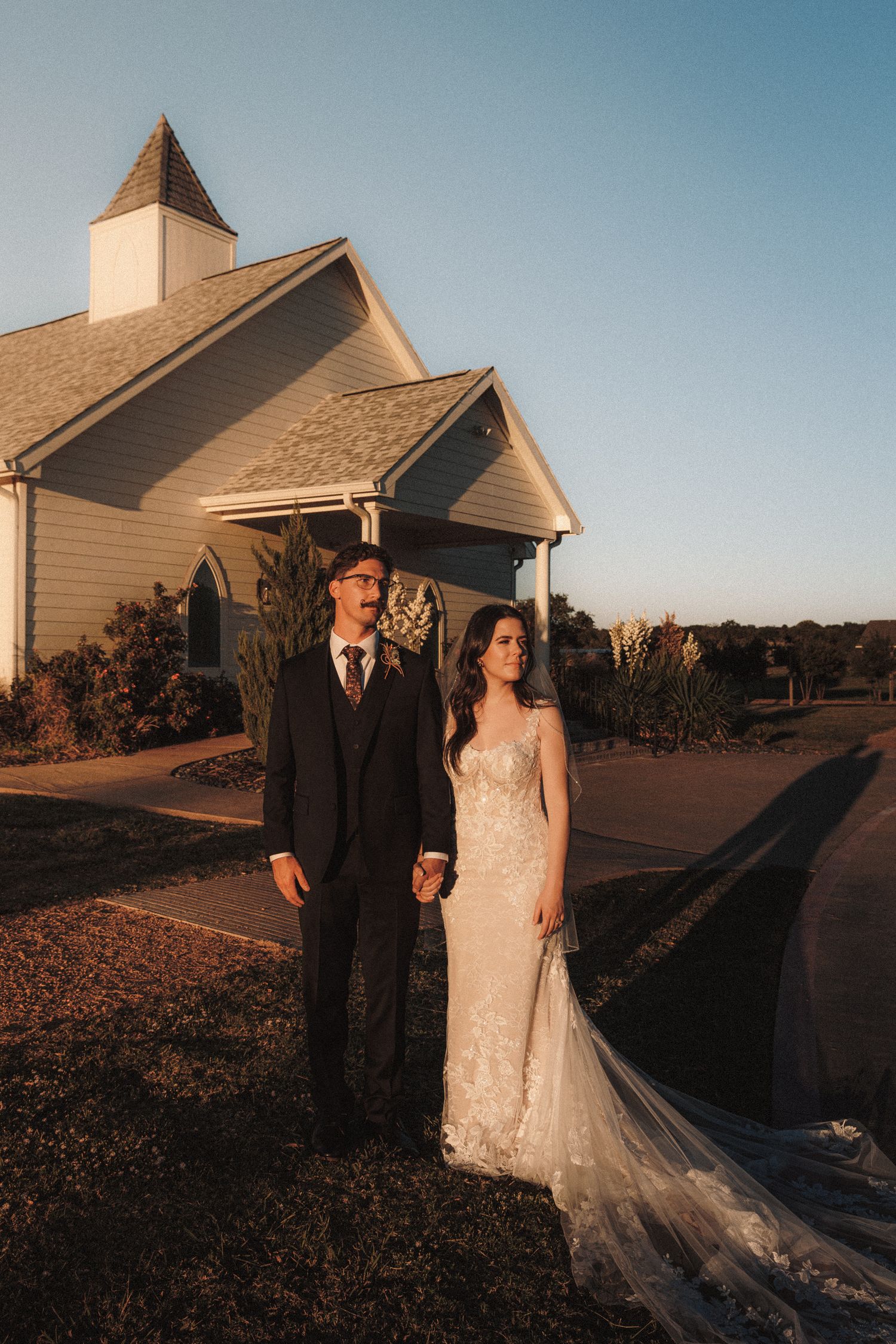 Bride and groom holding hands in golden sunset light outside charming white chapel wedding venue.