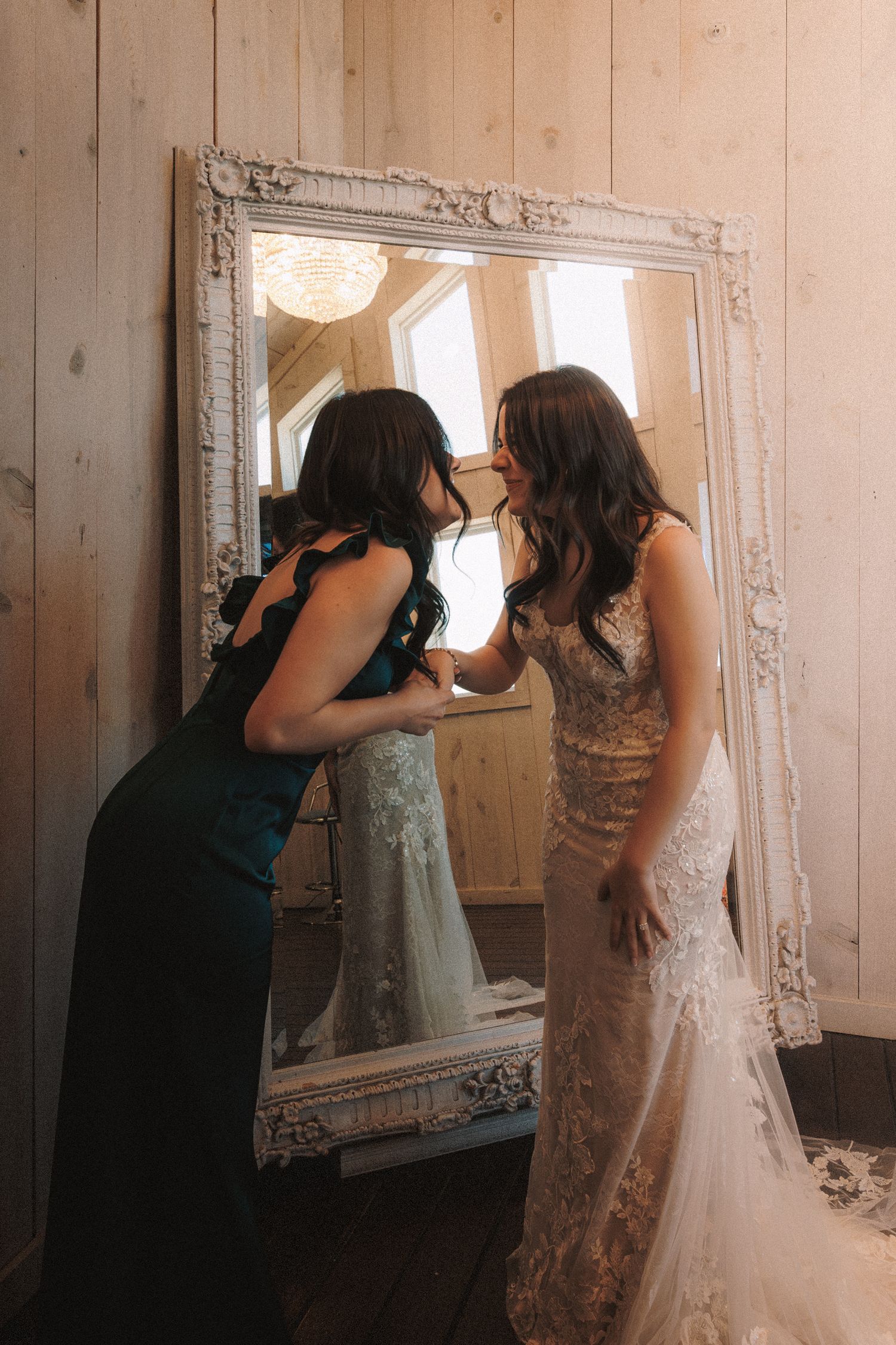Bridesmaid in teal dress leaning in close to the bride near an ornate white mirror.