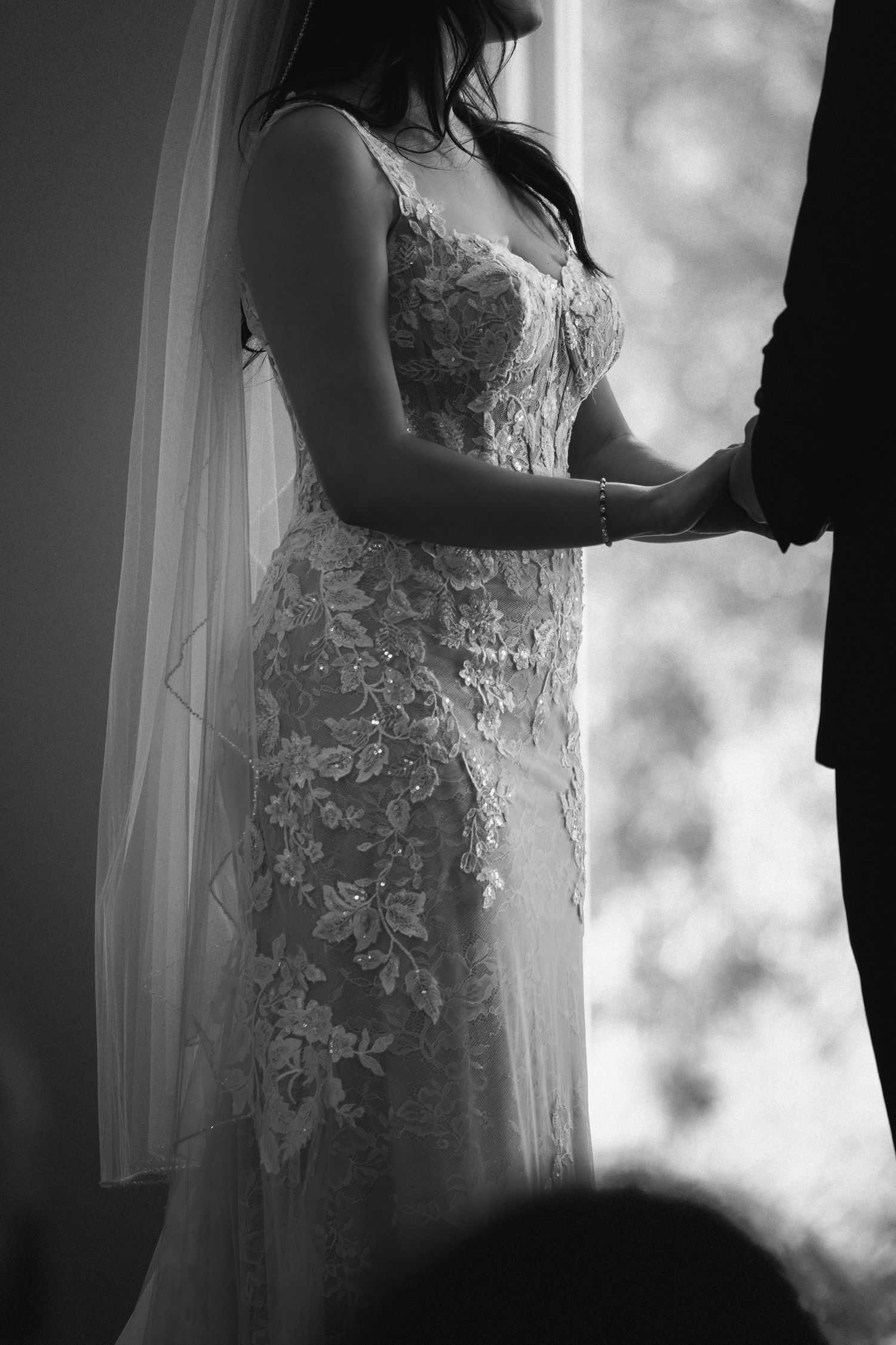 Black and white photo of bride in lace wedding gown holding hands with groom near a bright window.