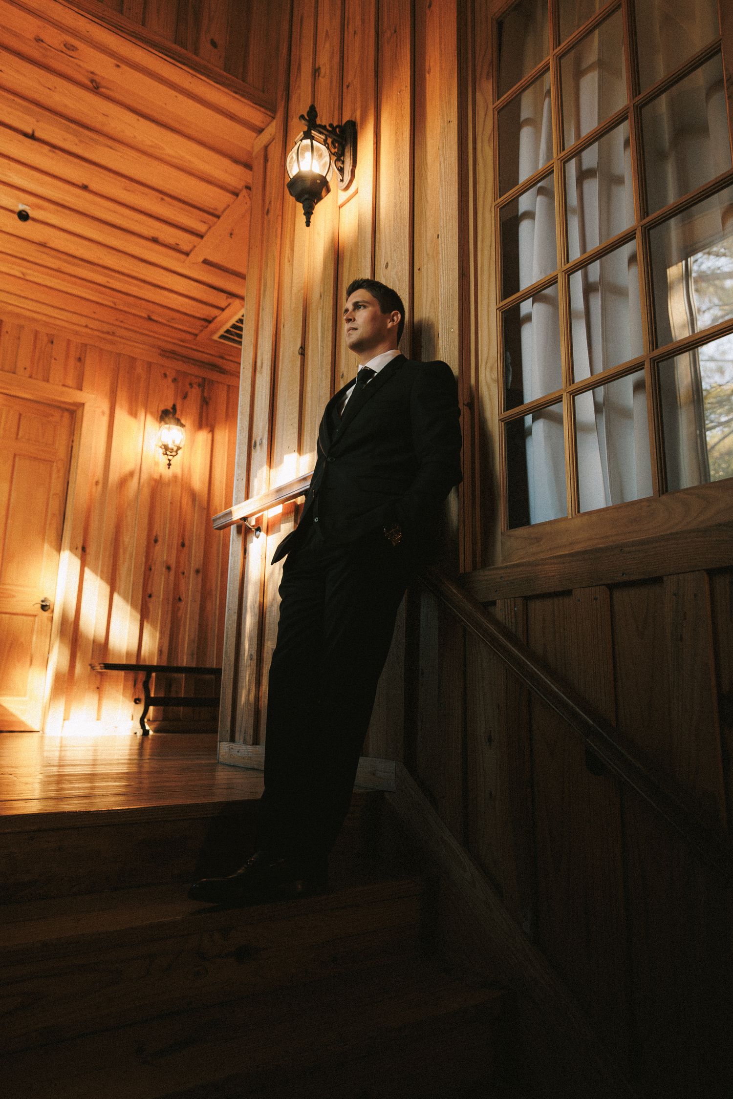Groom in dark suit standing on wooden staircase beside window in warmly lit rustic venue interior.