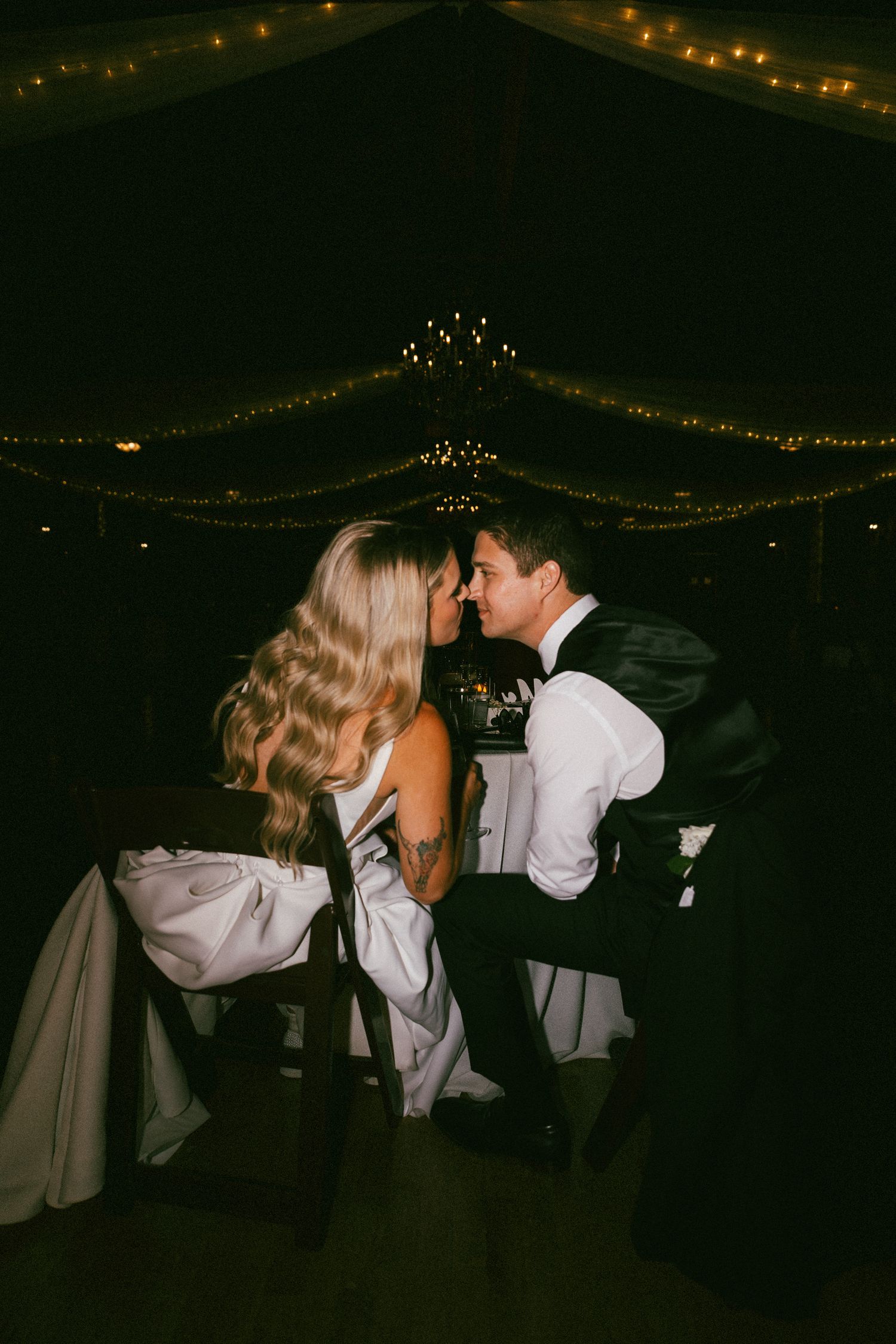 Bride and groom share a tender moment at their sweetheart table under warm string lights at evening reception.