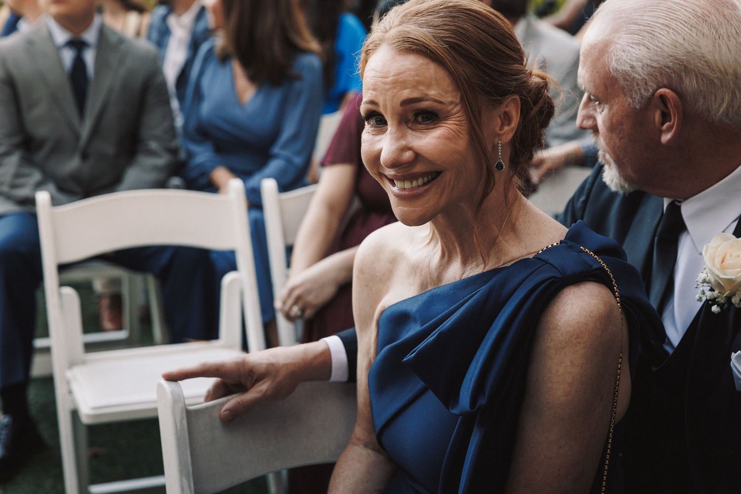 A smiling woman in a blue one-shoulder dress sits at an outdoor wedding ceremony.