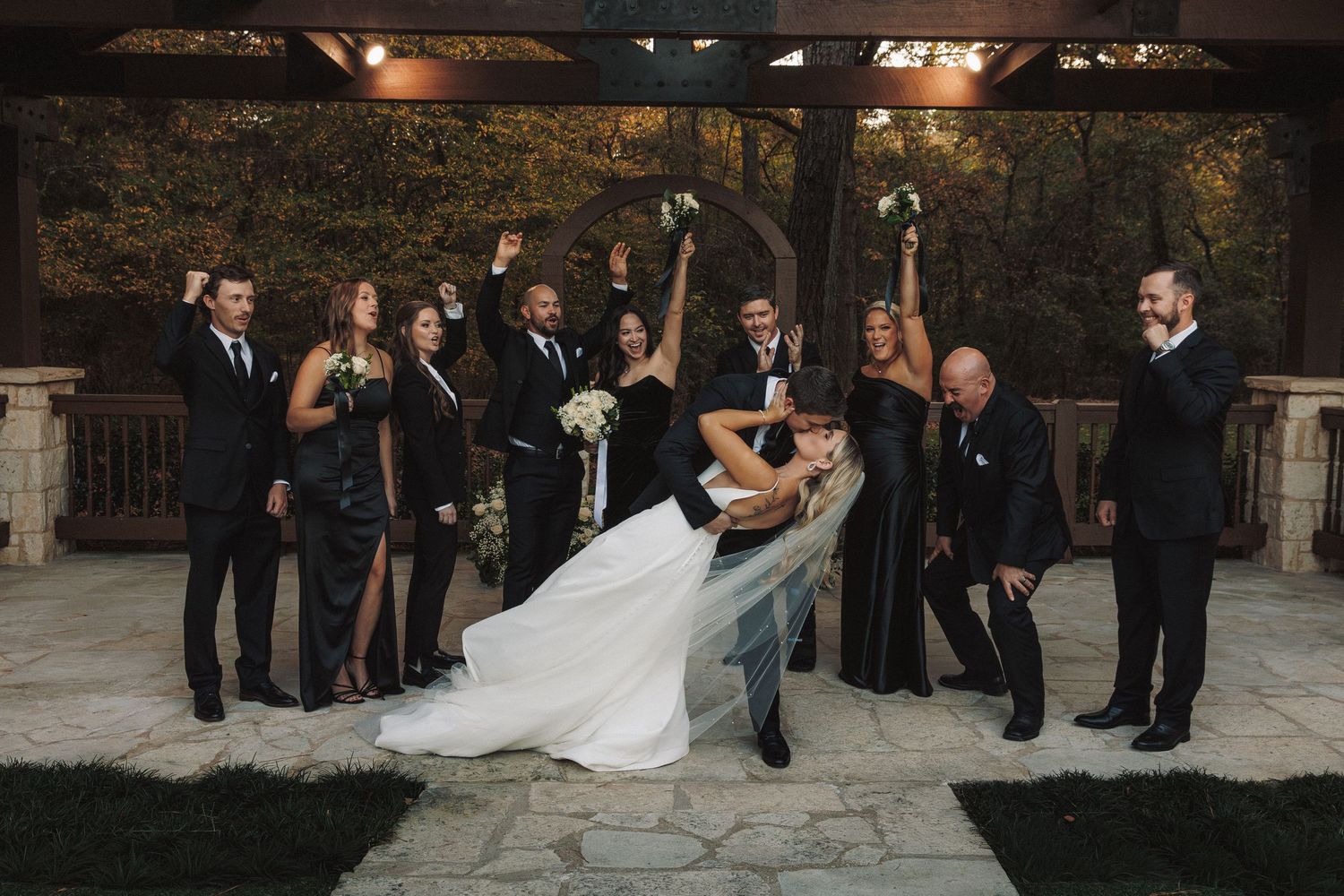 Wedding party in black attire cheers as groom dips bride under wooden outdoor pavilion.