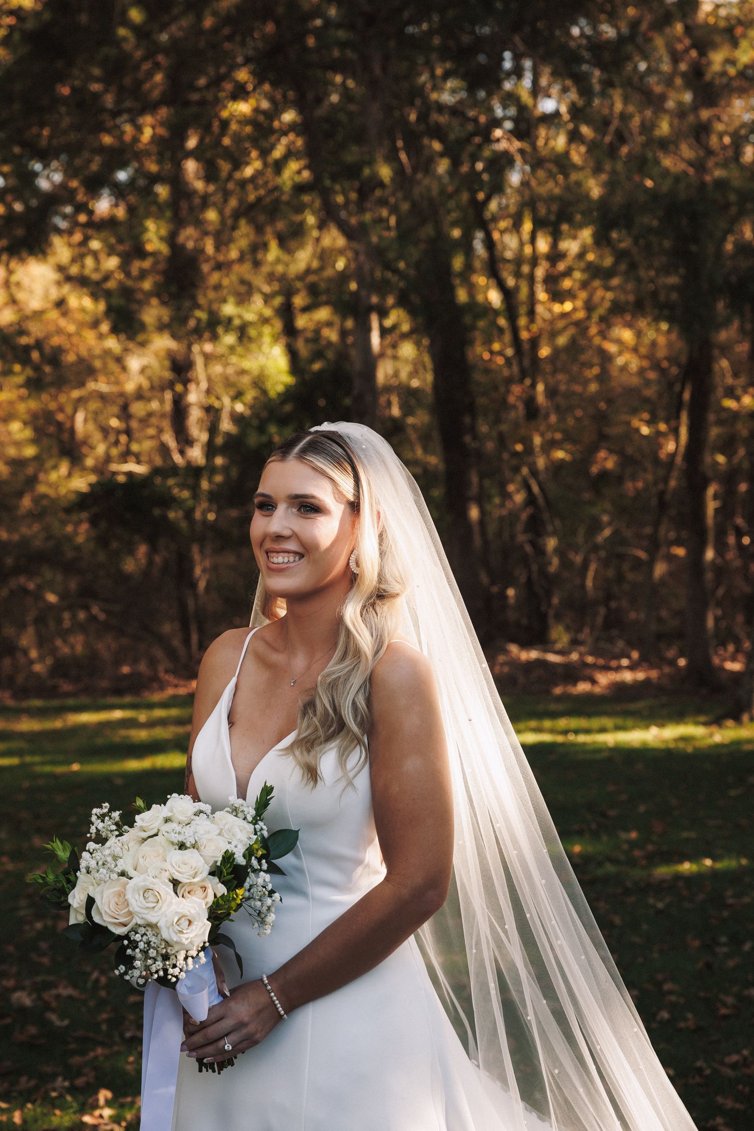 Beautiful bride in white gown and veil holding white rose bouquet smiling in autumn forest.