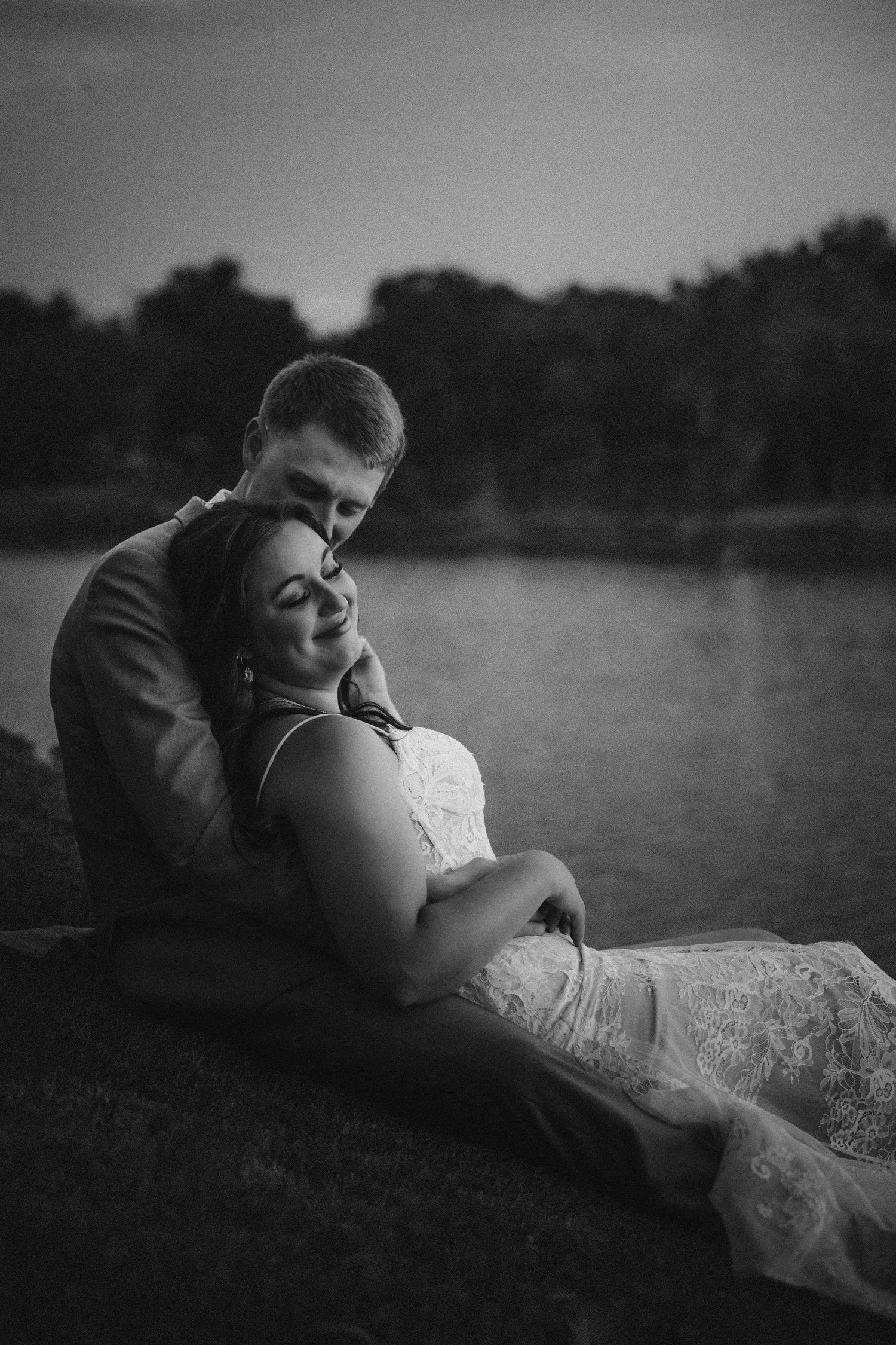 A romantic black and white wedding photo of a couple embracing near a serene lake at dusk.