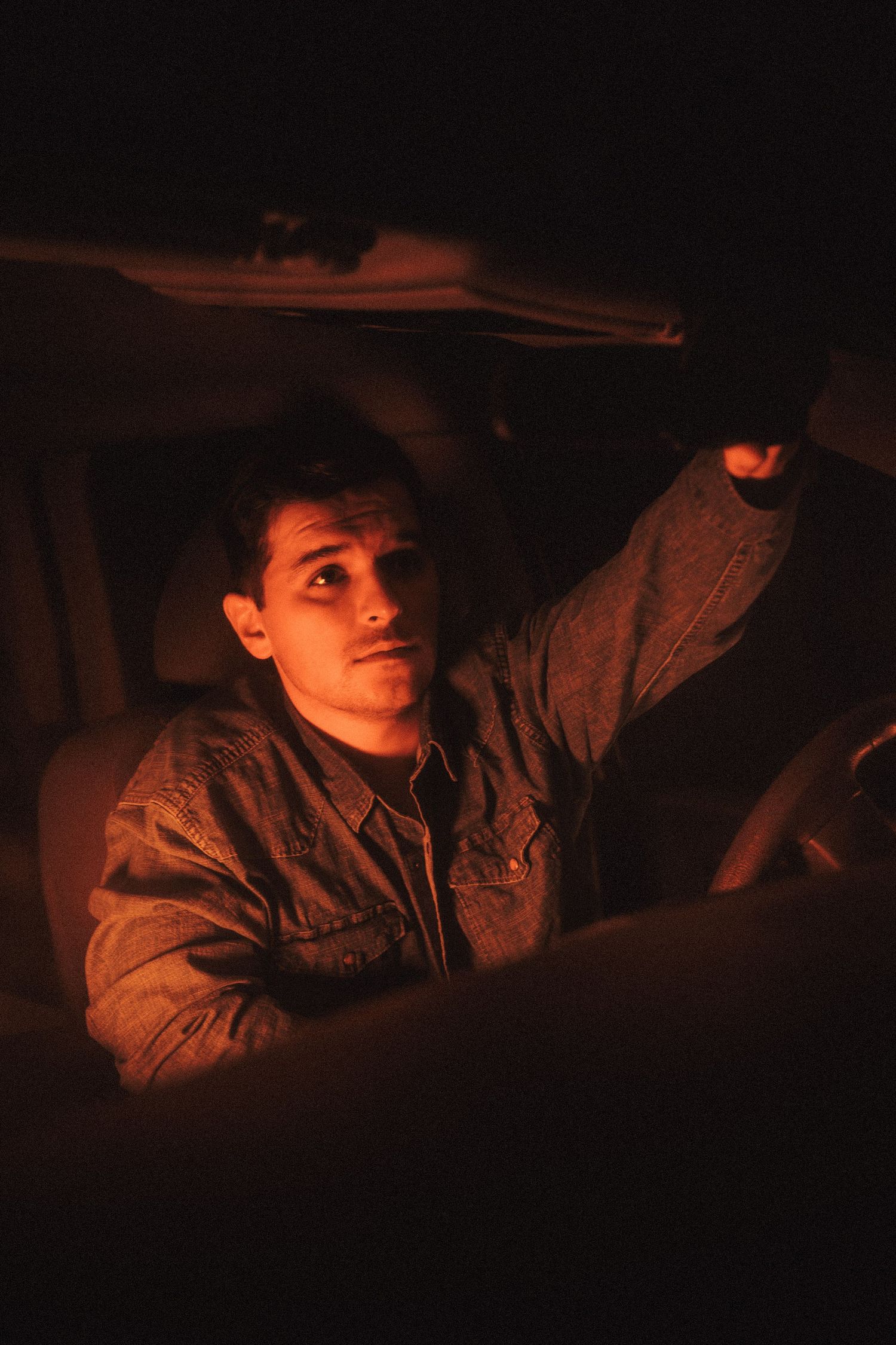 Figure illuminated by warm orange light sits in dark vehicle interior wearing brown jacket.