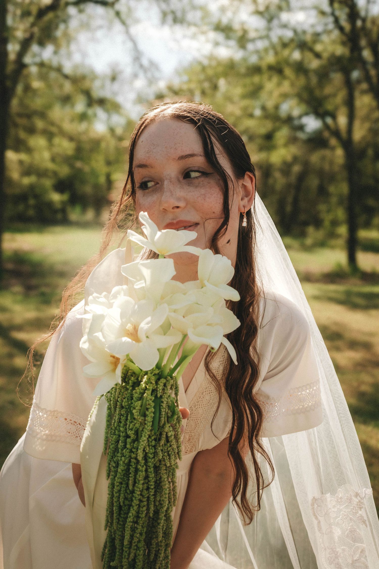 Bride in flowing veil holds elegant white lily bouquet in sunlit garden setting during outdoor wedding photoshoot.