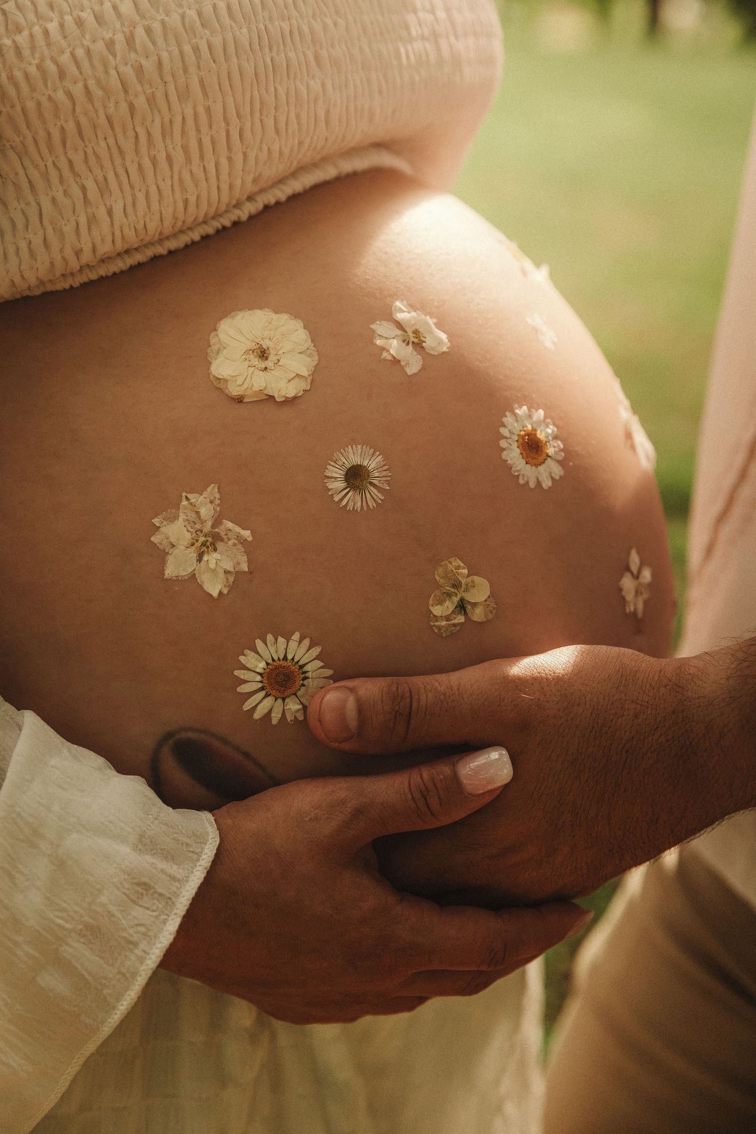 Small dried flowers delicately arranged against skin with hands on a pregnant belly in a series of soft, warm-toned photographs.