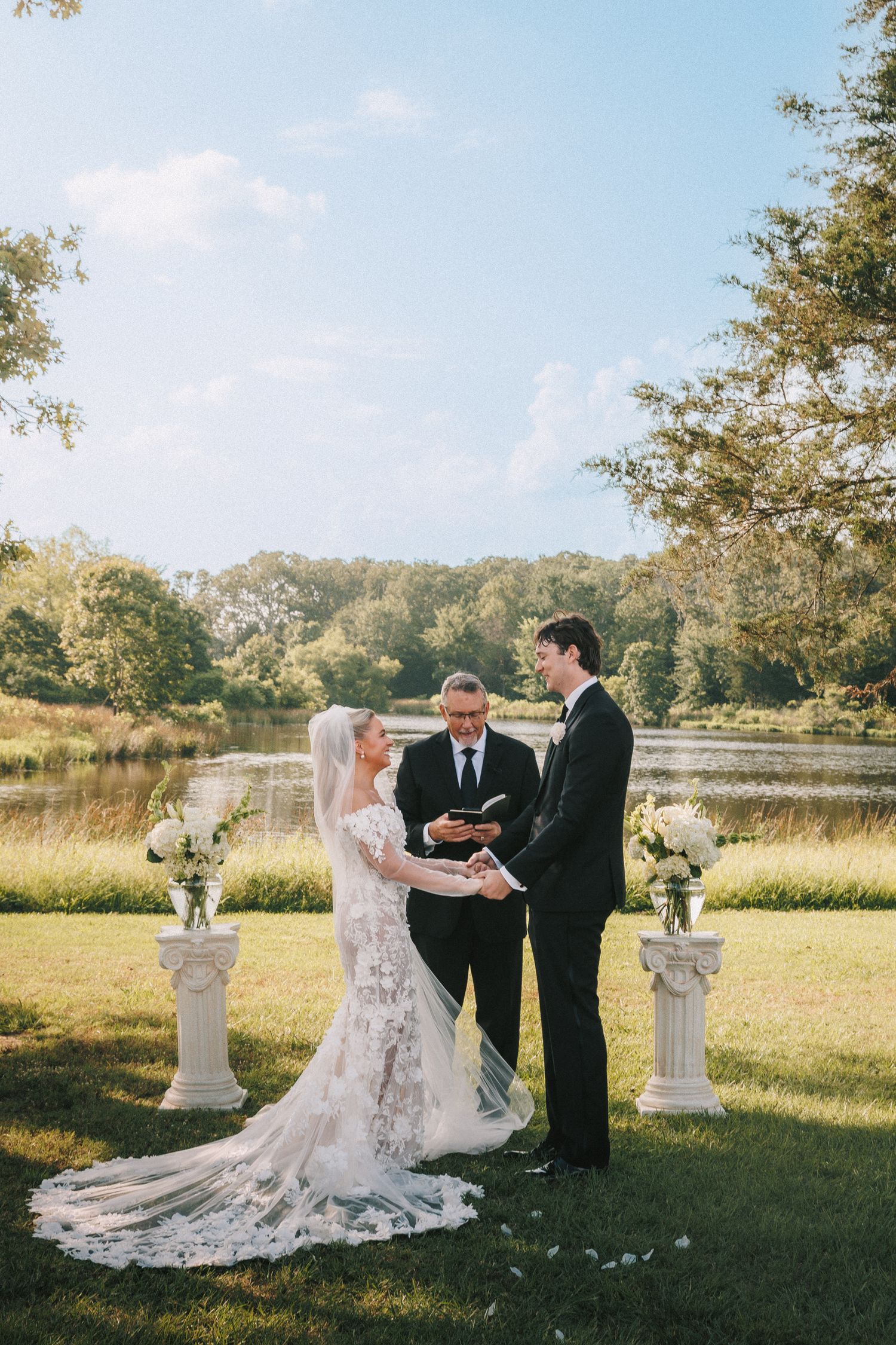 A wedding ceremony takes place outdoors by a lake with decorative white pillars and floral arrangements.