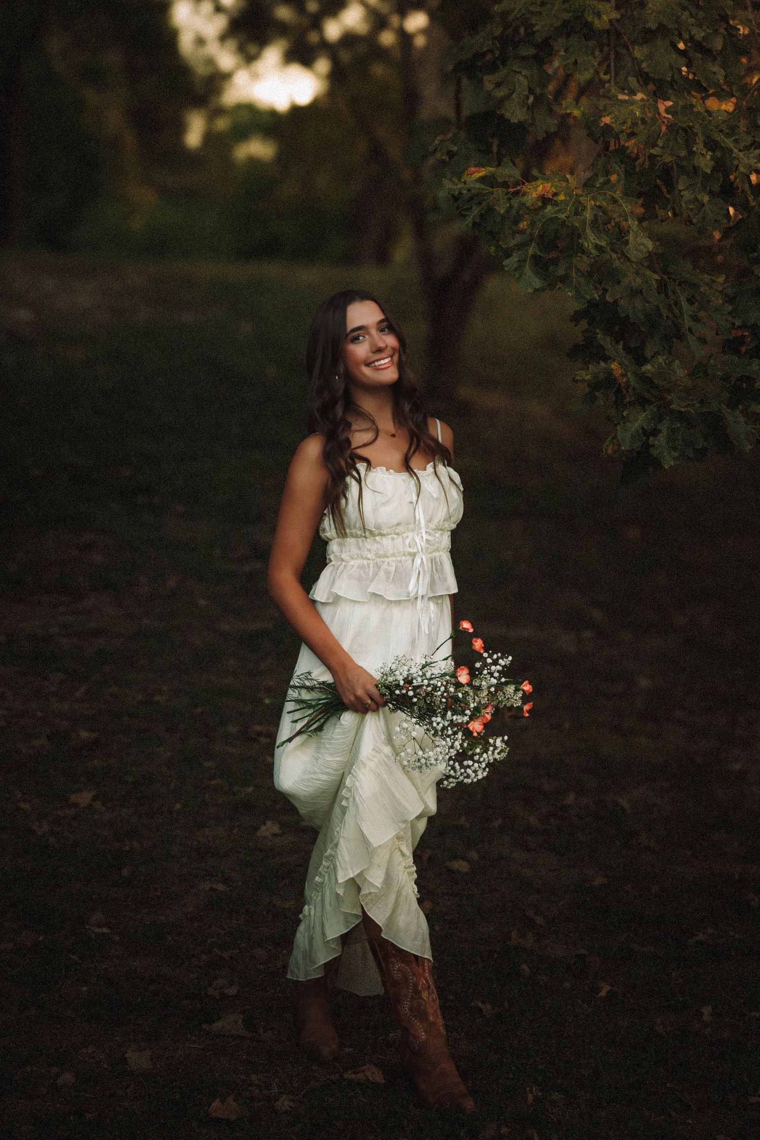 A person in a white dress and cowboy boots poses in a moody forest setting during golden hour.