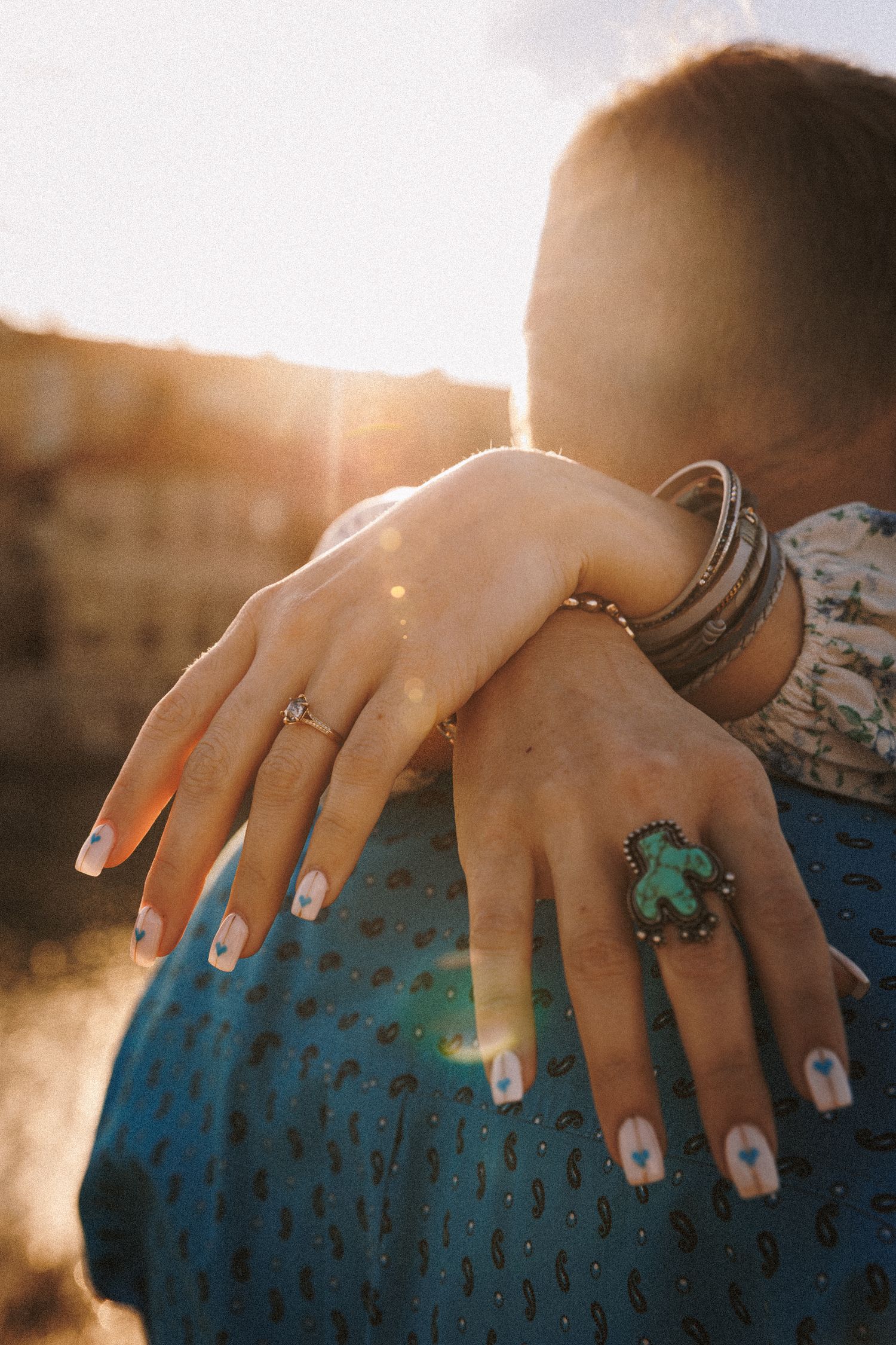 Close-up of hands with white nail polish and turquoise statement ring against sunlit backdrop.