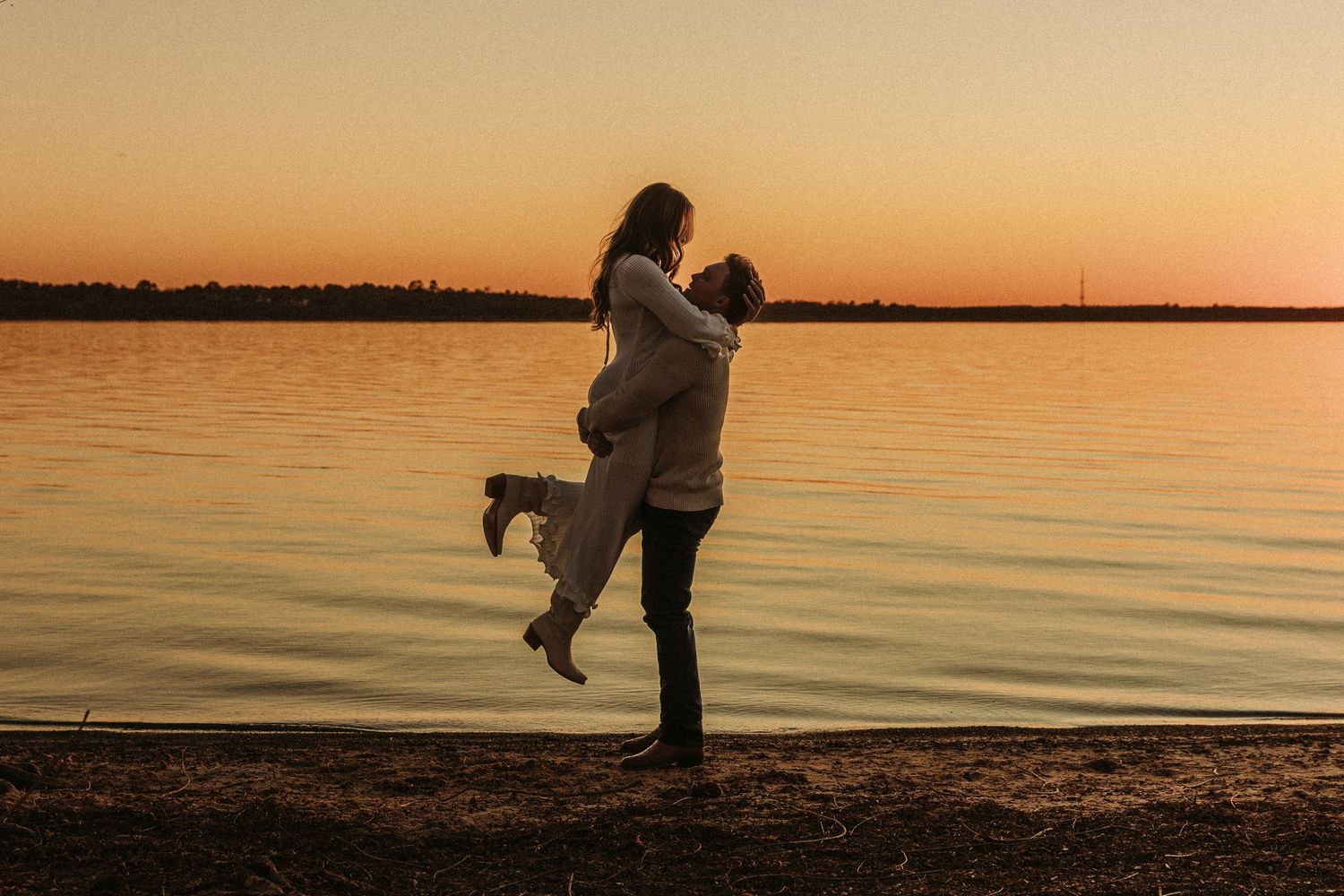 A romantic couple embraces on a beach during sunset with calm waters in the background.