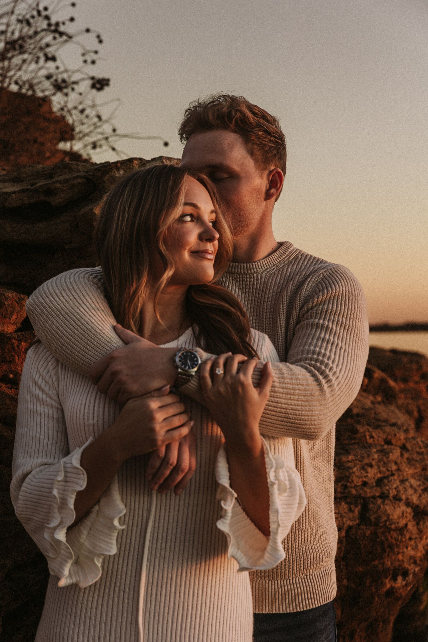 Romantic couple in matching sweaters embrace at sunset by rocky coastline.