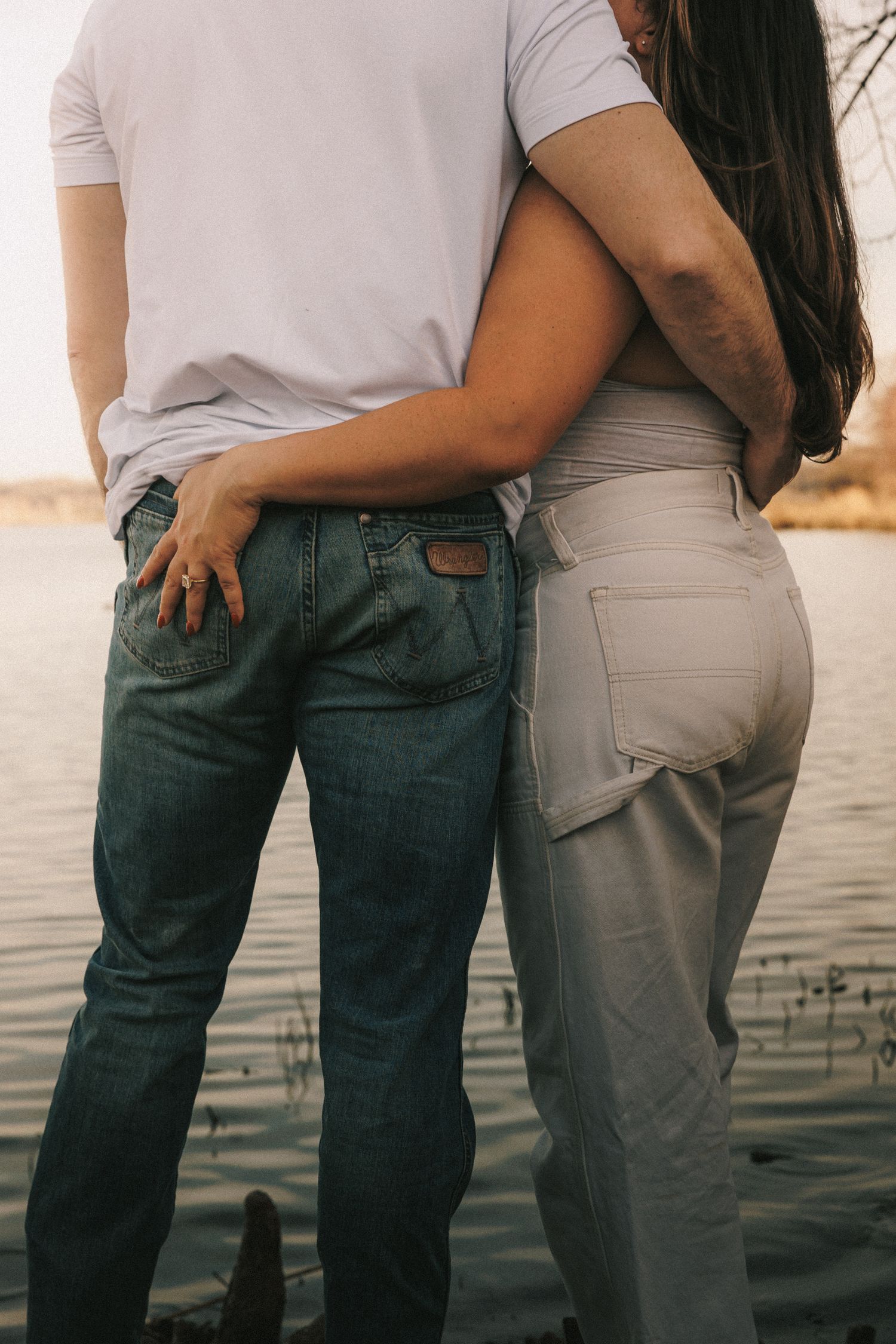 Close up of couple embracing on dock with water view at sunset, showing casual denim and white outfits.
