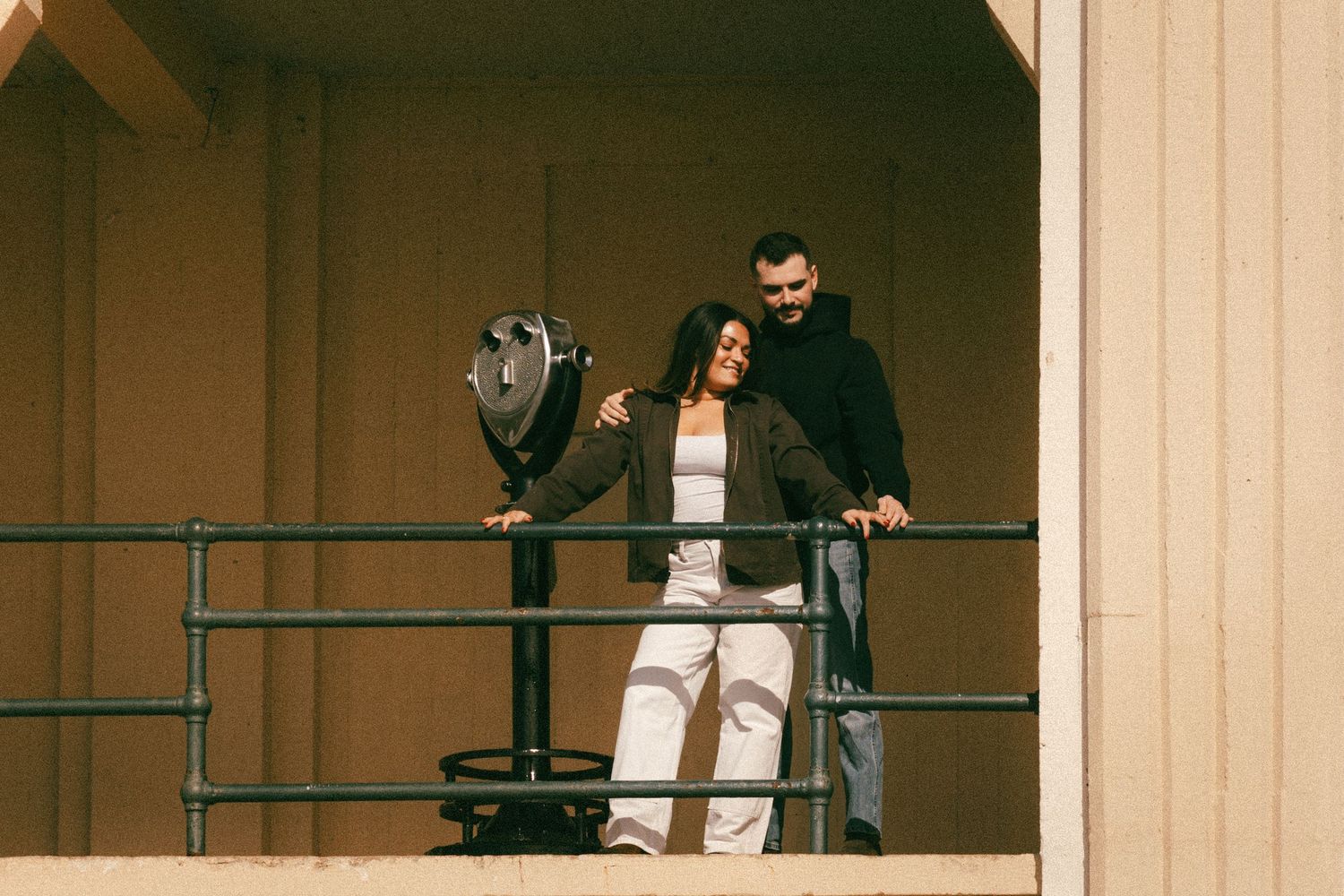 Two people leaning against a railing on a balcony in casual attire during golden hour.