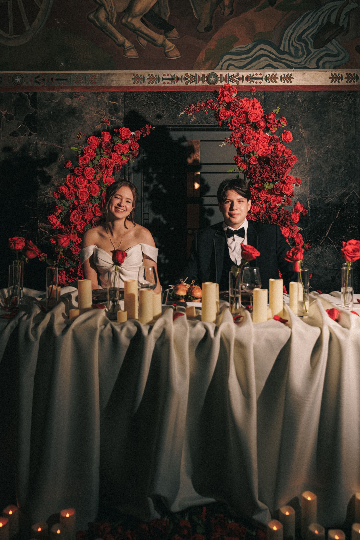 Bride and groom seated at sweetheart table framed by lush red rose floral arch with candles.
