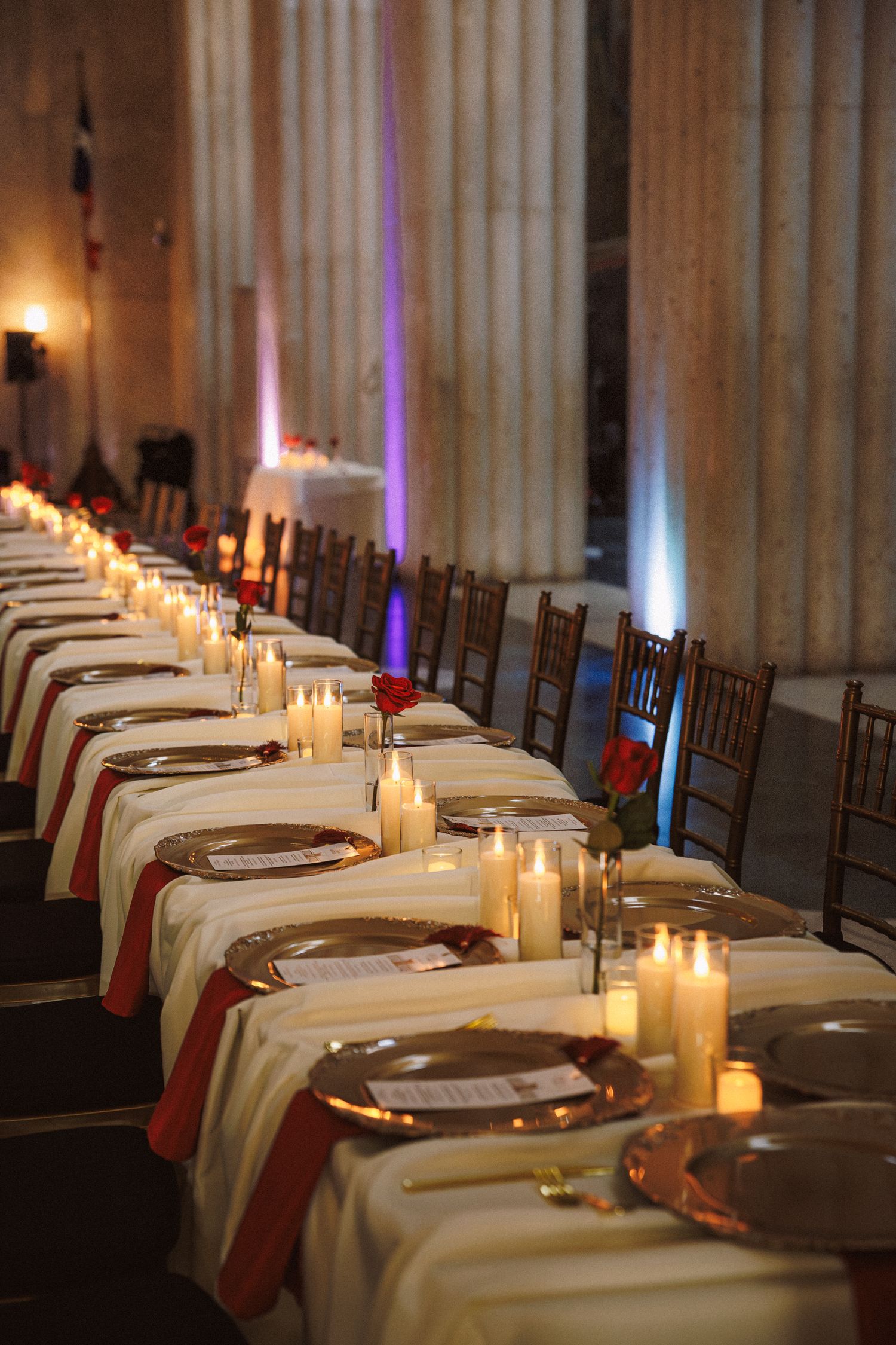 Elegant banquet hall with long candlelit table, gold chargers, red napkins, and tall columns in warm ambient lighting.