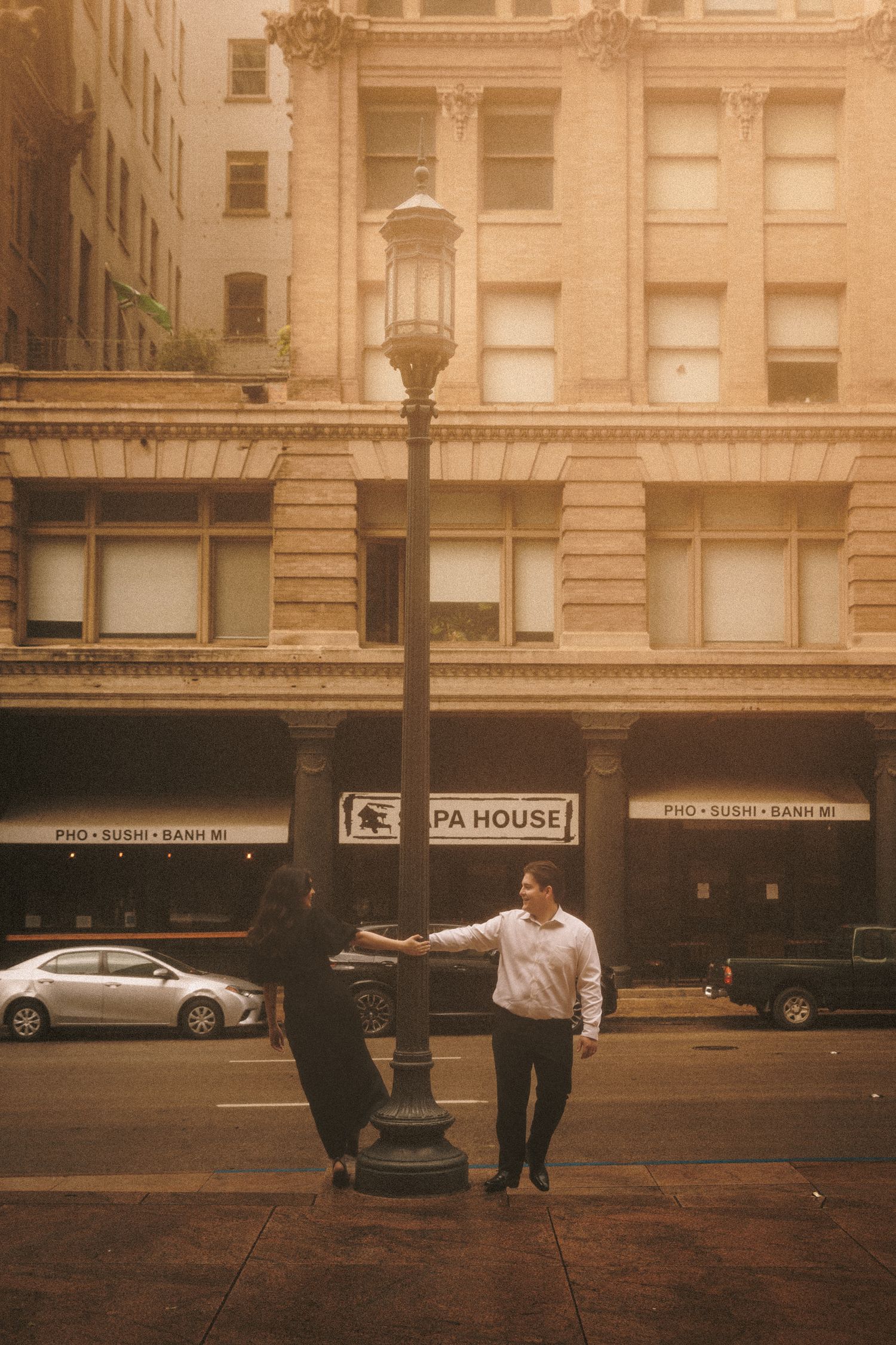 Couple leaning against a street lamp on a foggy urban sidewalk with vintage buildings and a pho restaurant in the background.