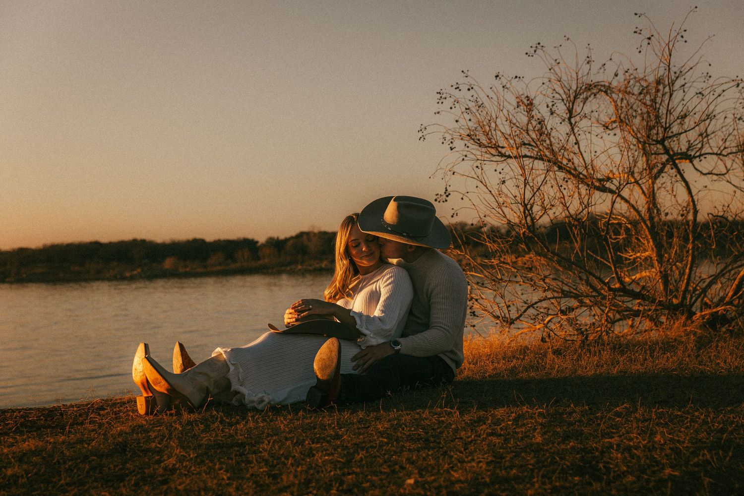 Dallas, Texas Engagement Photographer A couple relaxes by a tranquil lake at sunset, sitting on the grassy shore in warm golden light.