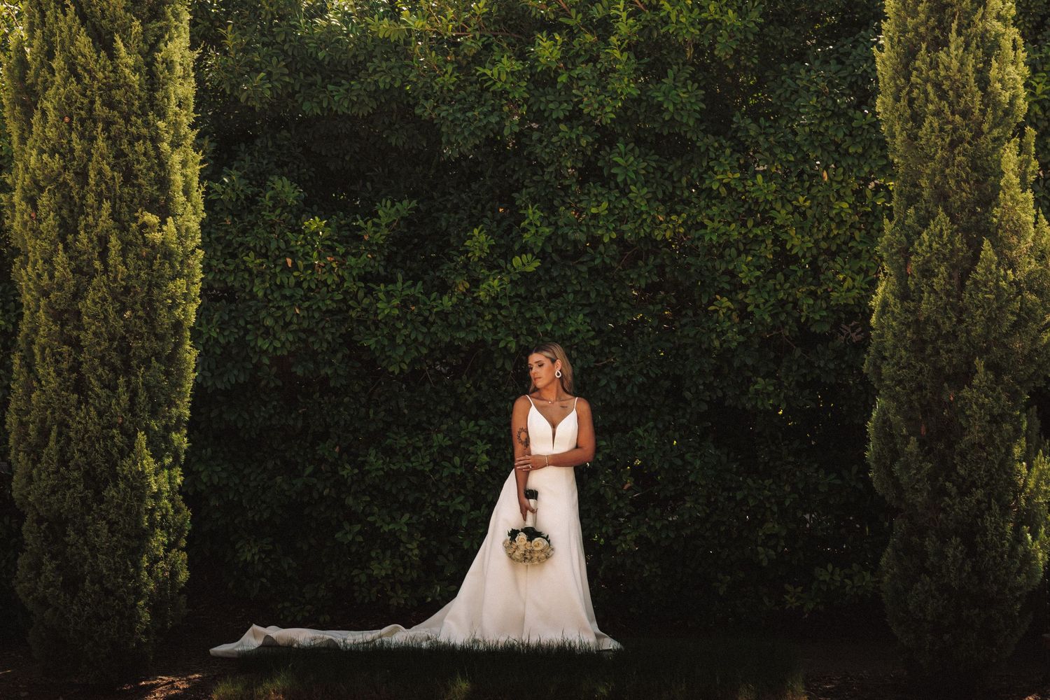 A person in a white wedding dress stands between tall cypress trees in a garden setting at dusk.