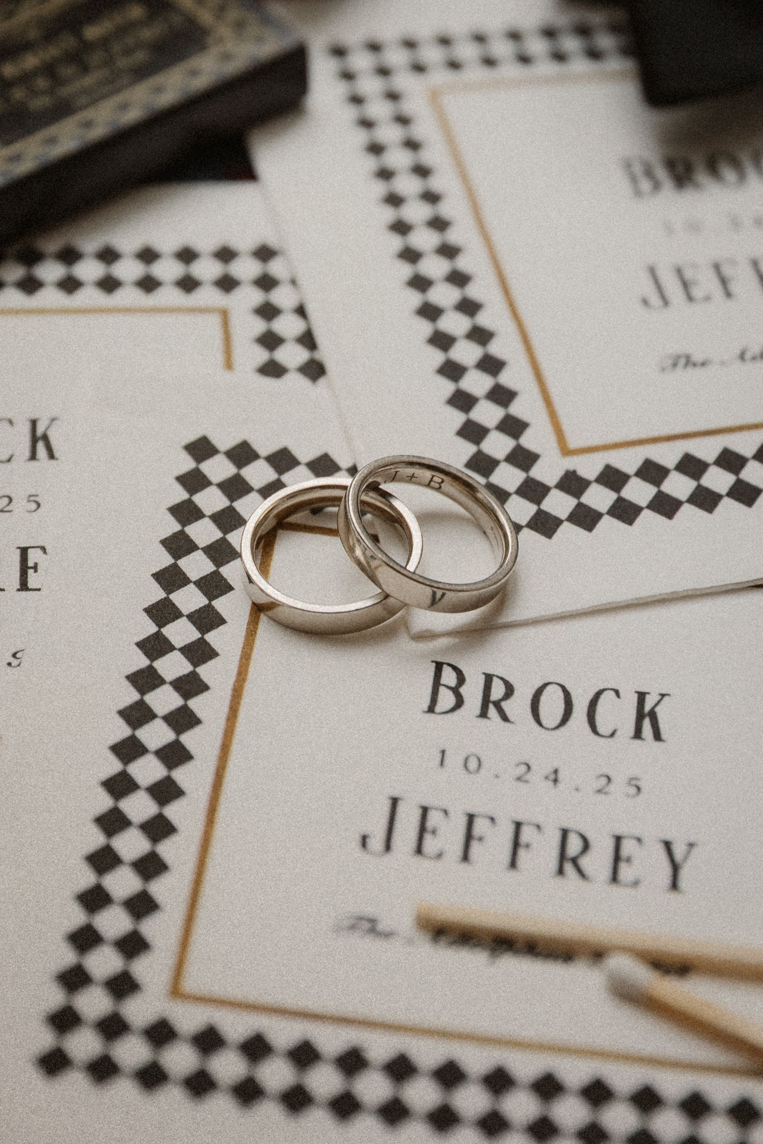 Two silver wedding bands rest on elegant wedding invitations for Brock and Jeffrey dated 10-24-25 with checkered borders.