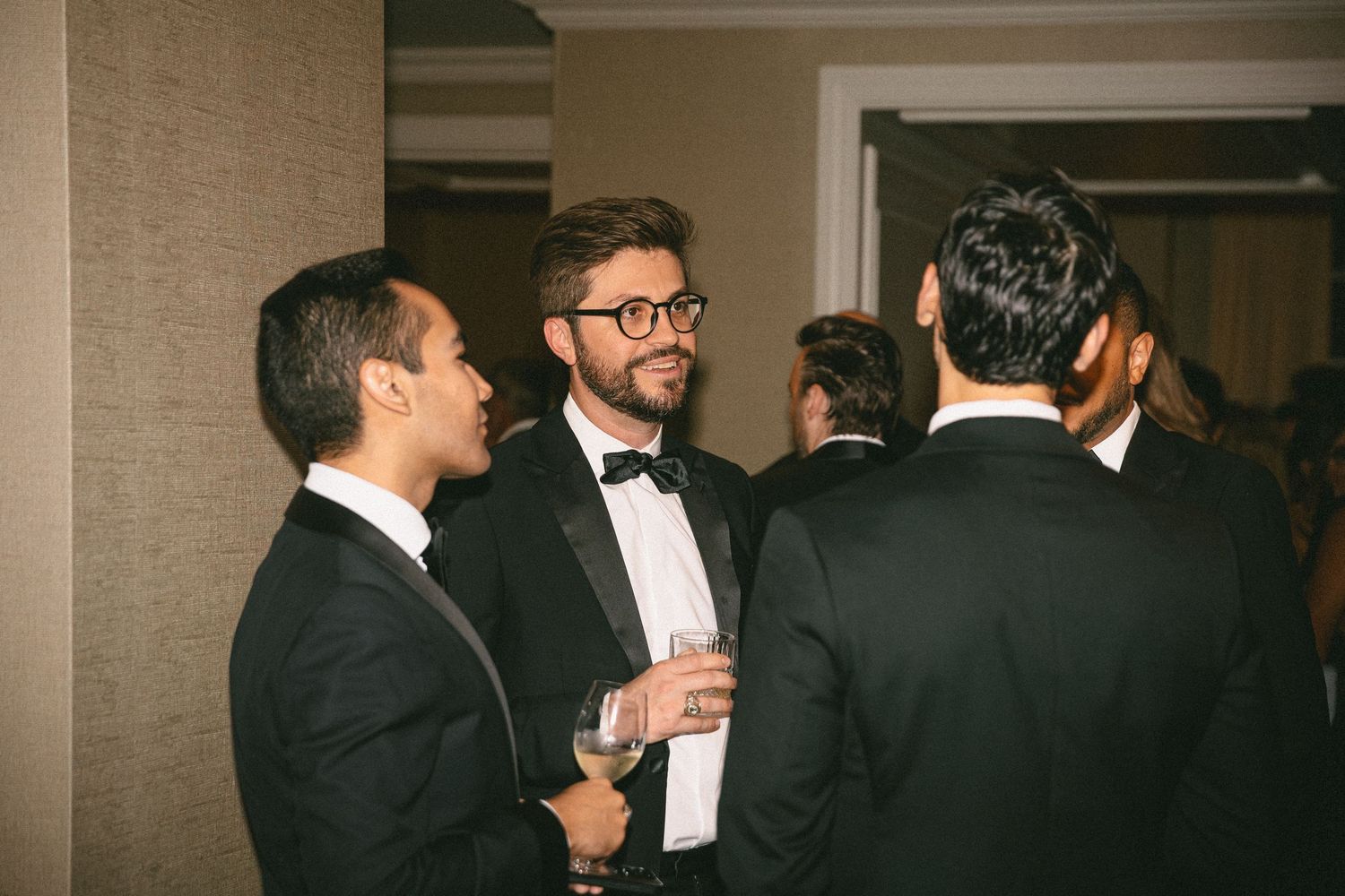Wedding guest in black tuxedo and glasses socializes with other formally dressed guests at reception cocktail hour.