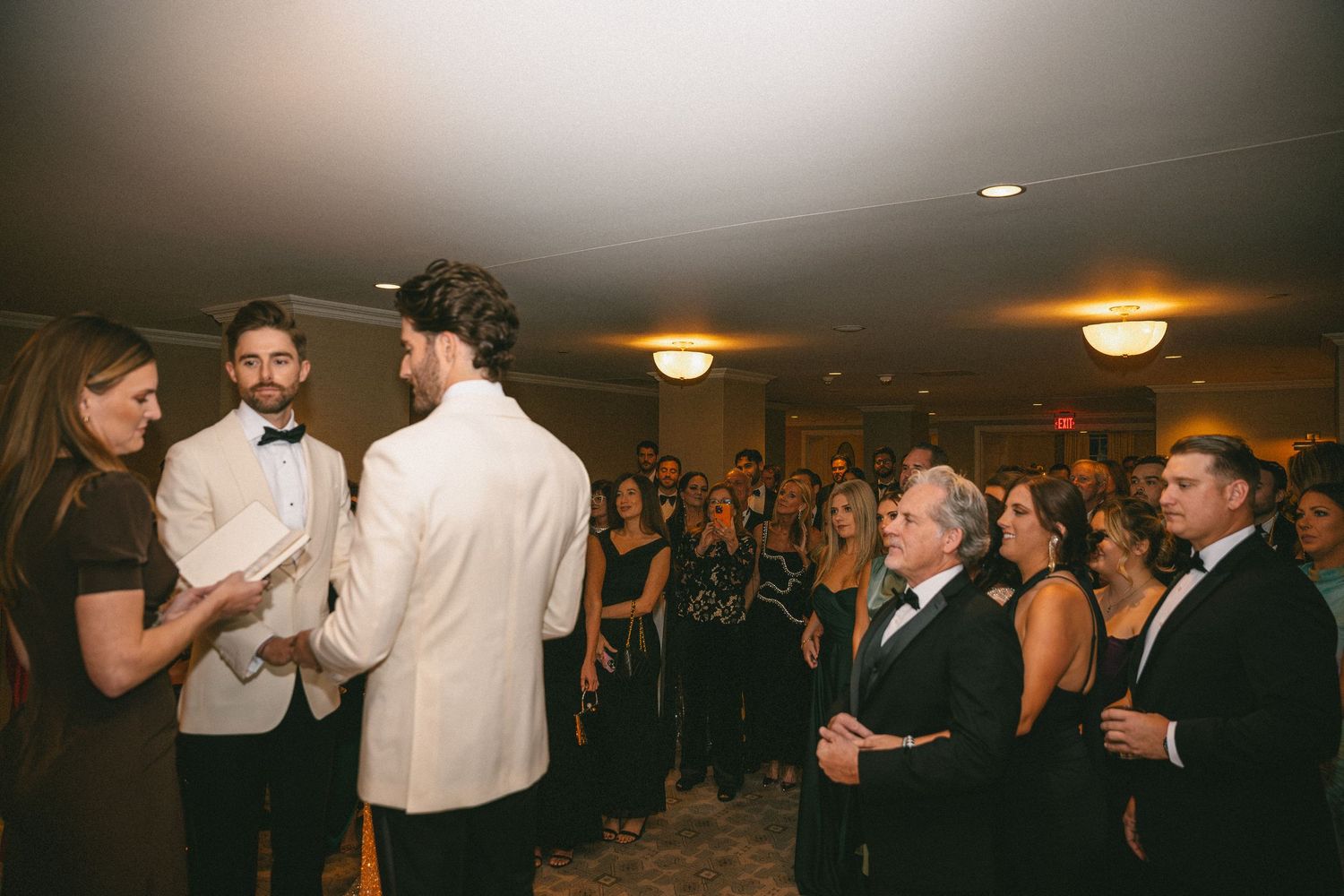Two grooms exchanging vows before an emotional crowd of wedding guests in a warmly lit ballroom ceremony.