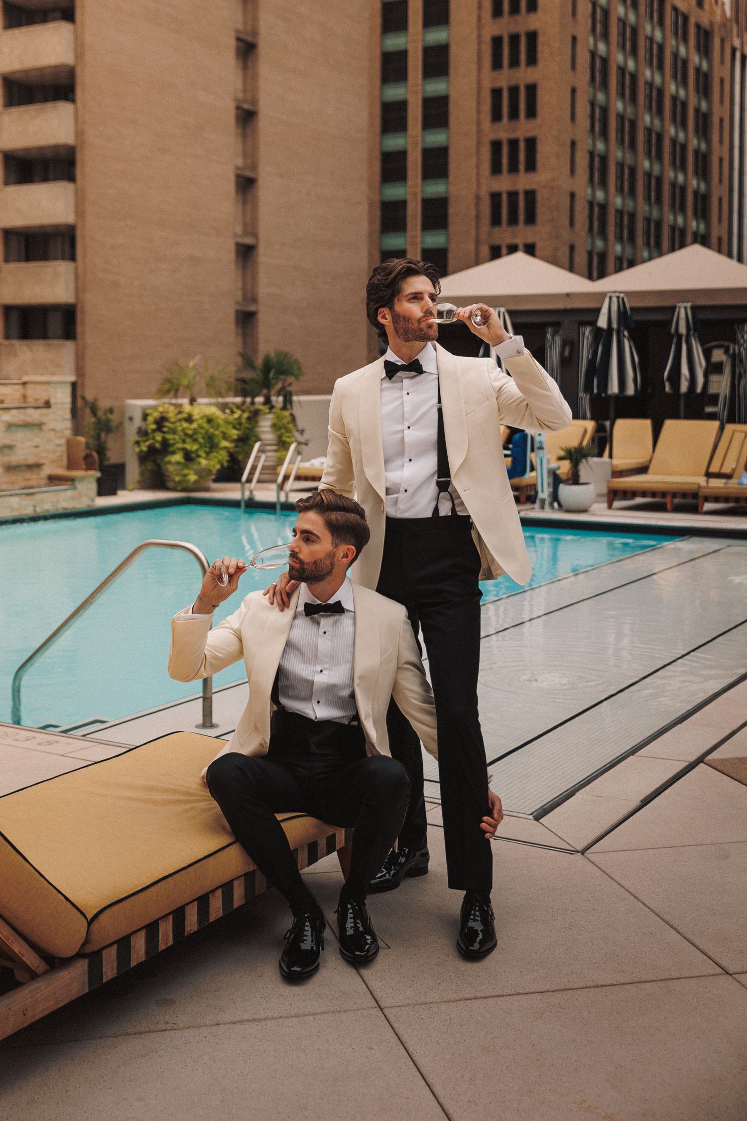 Two grooms in ivory tuxedos pose elegantly by a rooftop pool at their wedding.