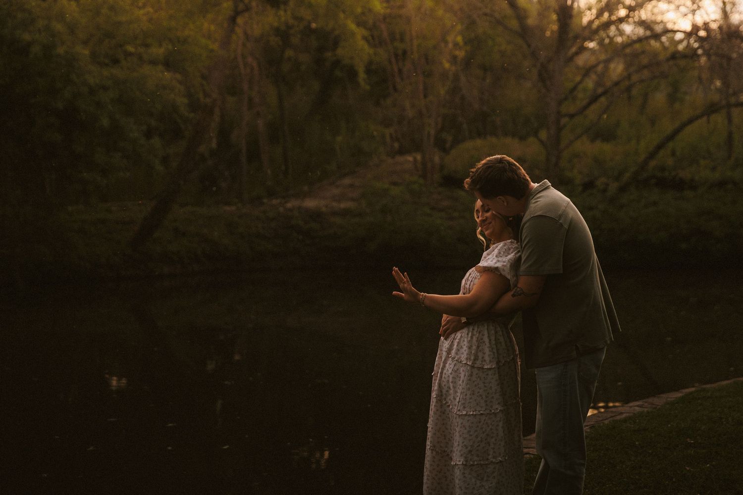 Couple standing together by a peaceful pond in soft golden evening light among trees.