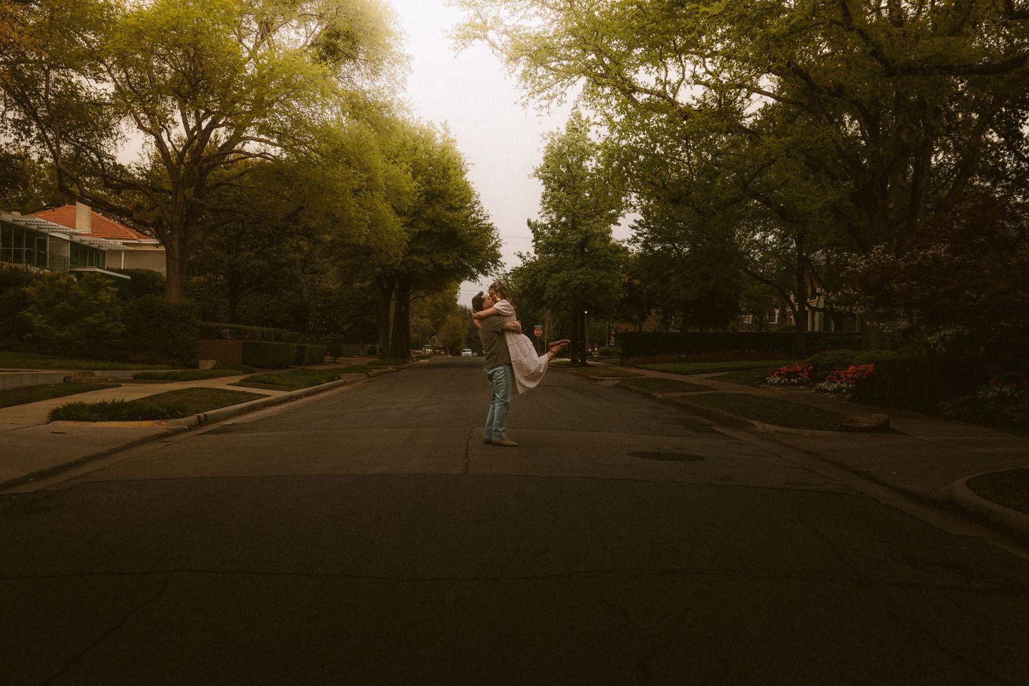 A couple embracing and spinning joyfully in the middle of a tree-lined residential street at dusk.