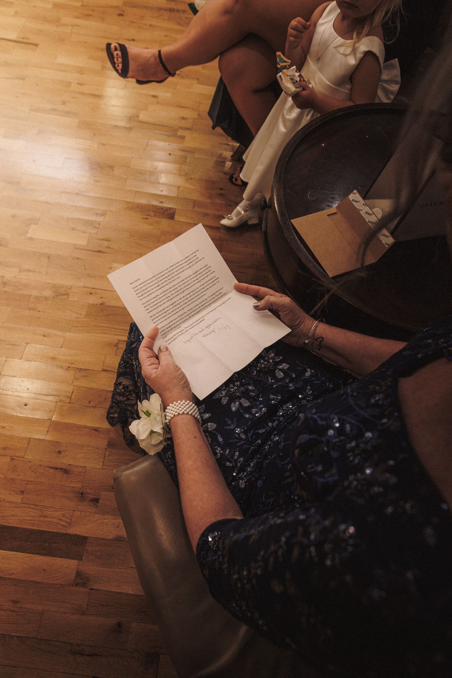 Woman in navy sequin dress reads wedding program while seated on wooden floor at elegant ceremony.