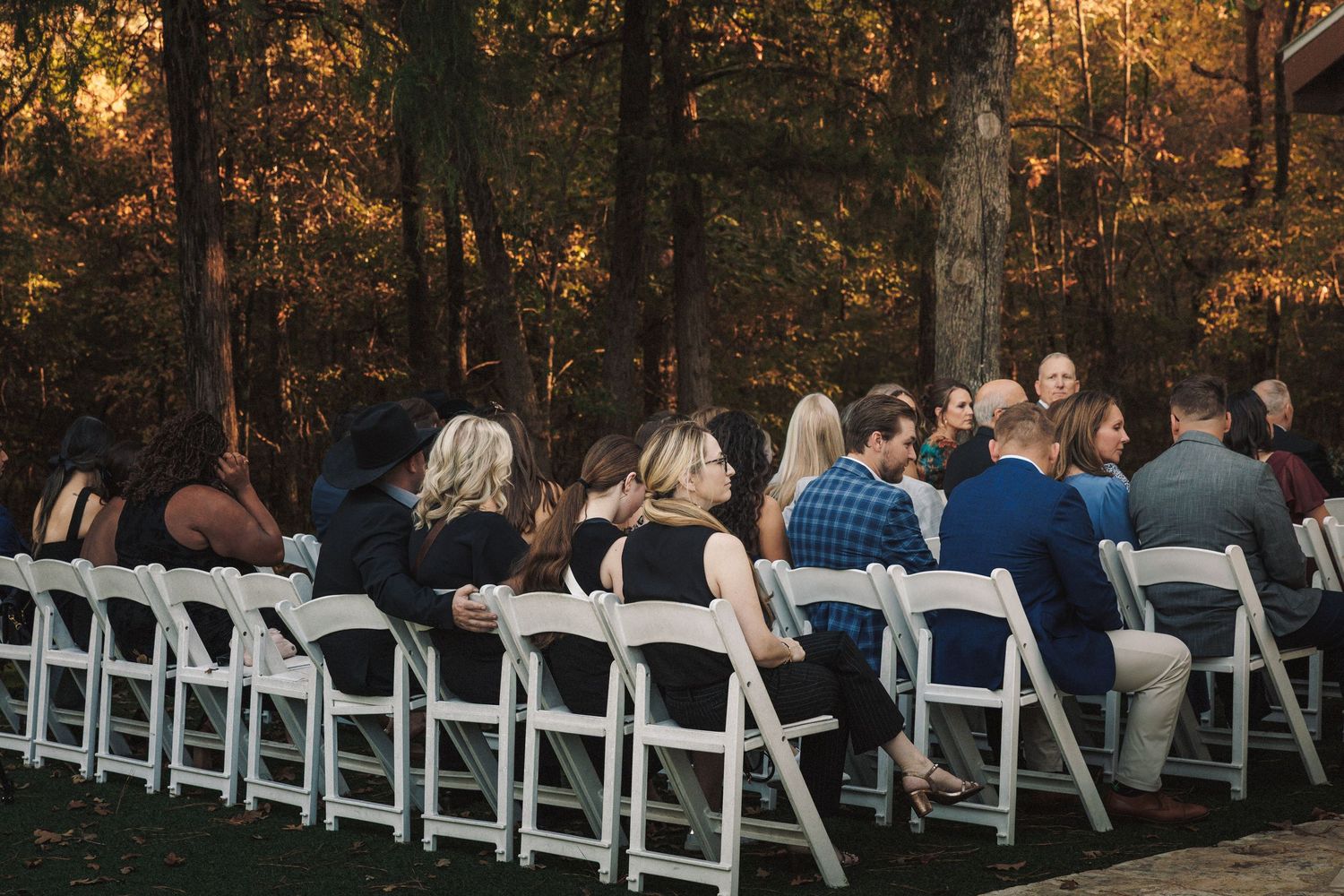 Wedding guests seated in white folding chairs at an outdoor ceremony surrounded by tall trees.
