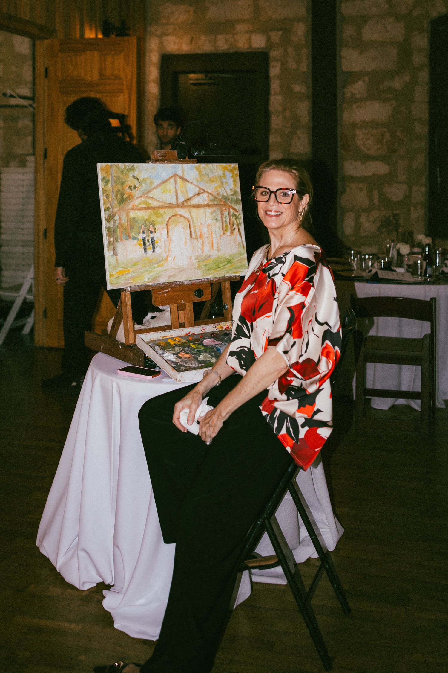 A smiling woman in floral top sits beside a live wedding painting on an easel in a rustic venue.