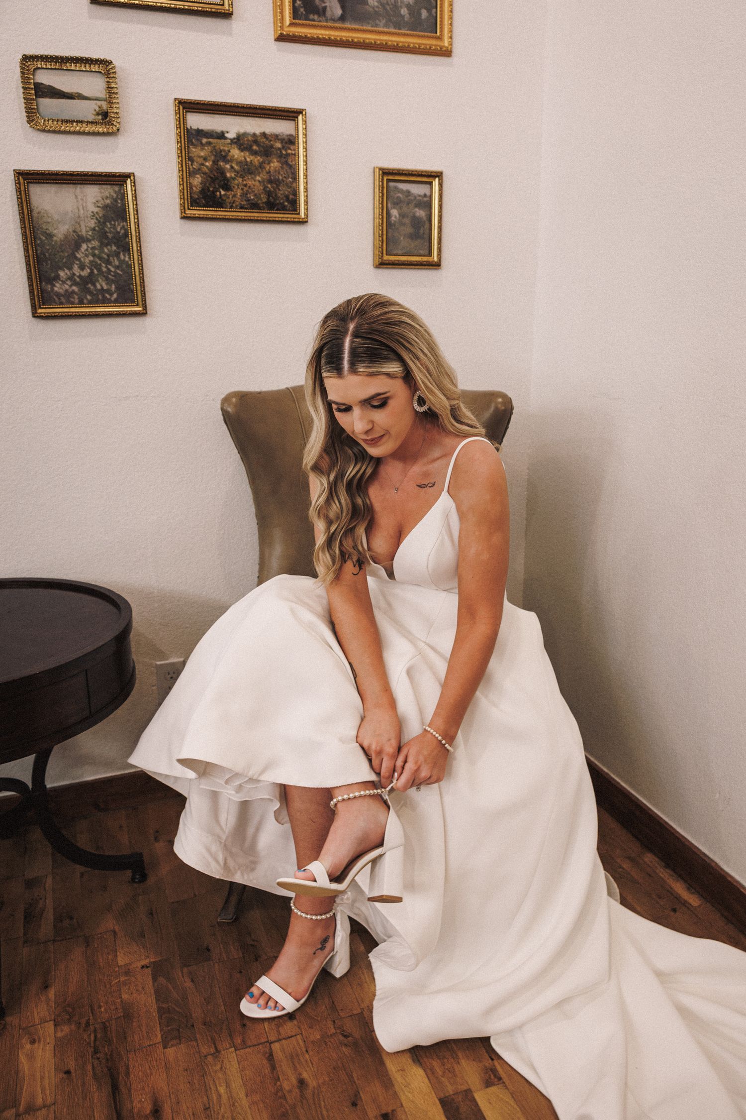 Bride in white gown adjusting her heeled sandal while seated in a chair before her wedding ceremony.