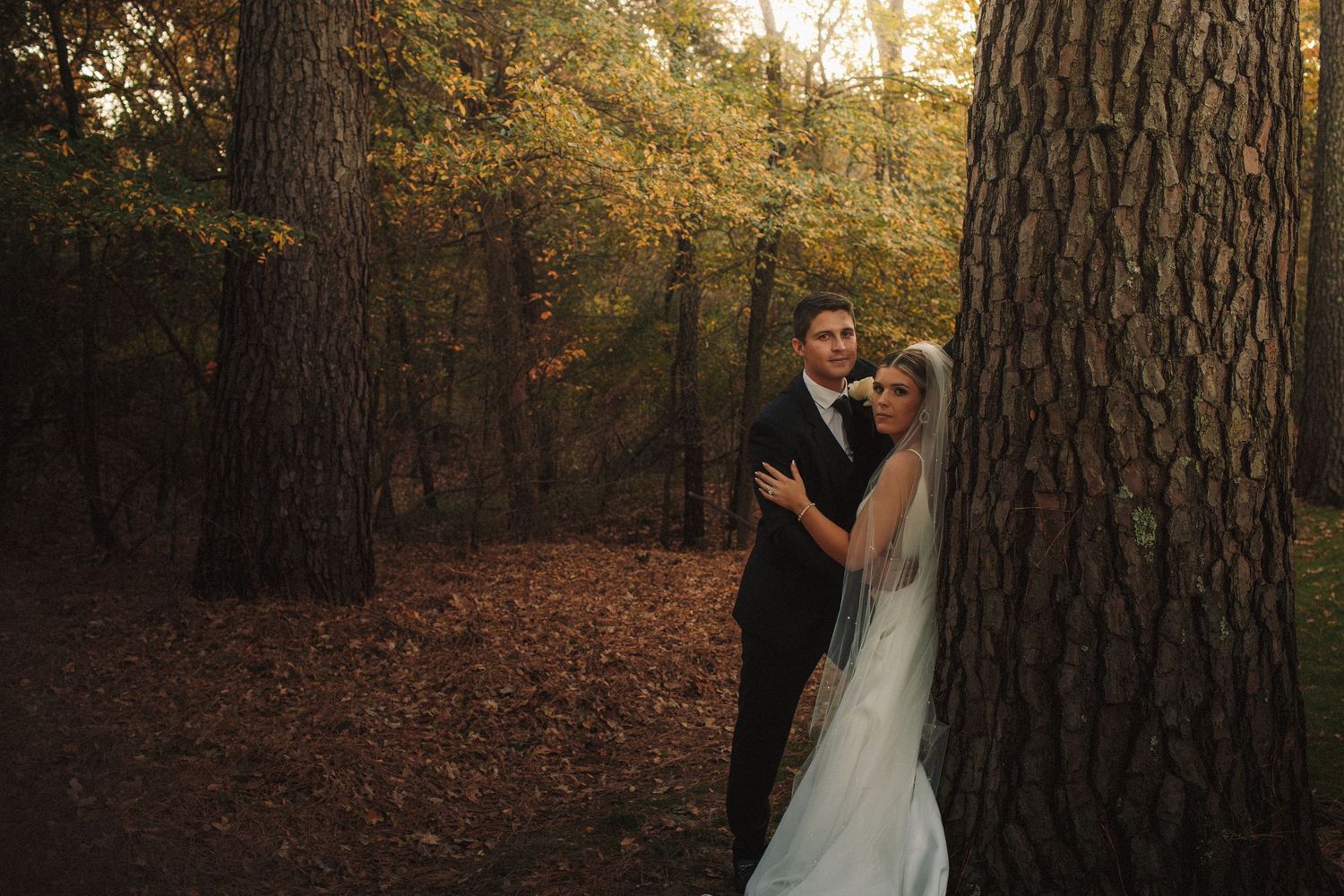 Bride and groom pose romantically beside a large tree trunk in a golden autumn forest wedding portrait.
