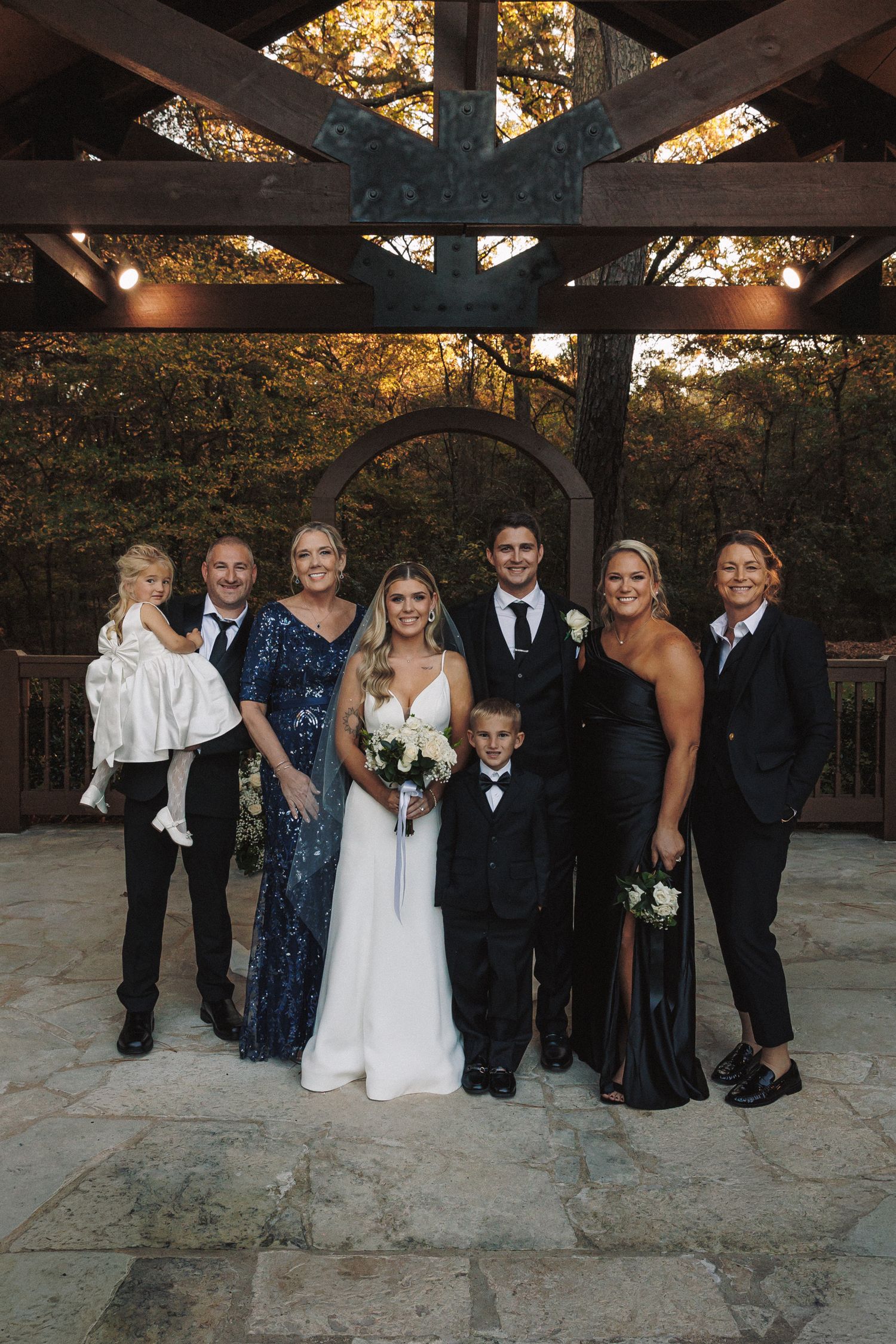 Wedding party family portrait under a rustic wooden pergola surrounded by colorful autumn foliage.