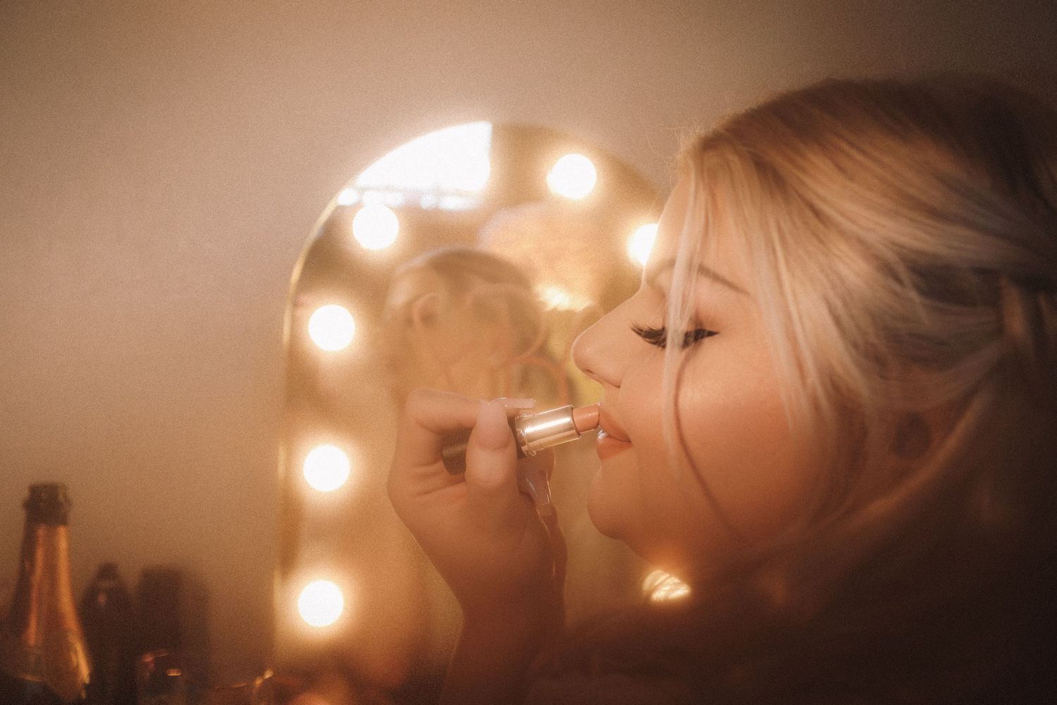 Close-up of blonde bride applying lipstick at glowing Hollywood vanity mirror with champagne nearby.