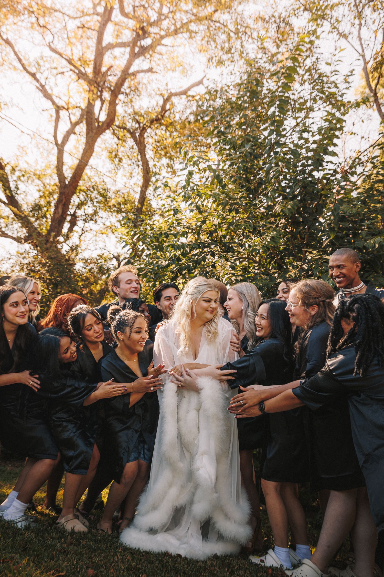 Bride in white fur stole surrounded by wedding party in black robes outdoors in golden sunlight.