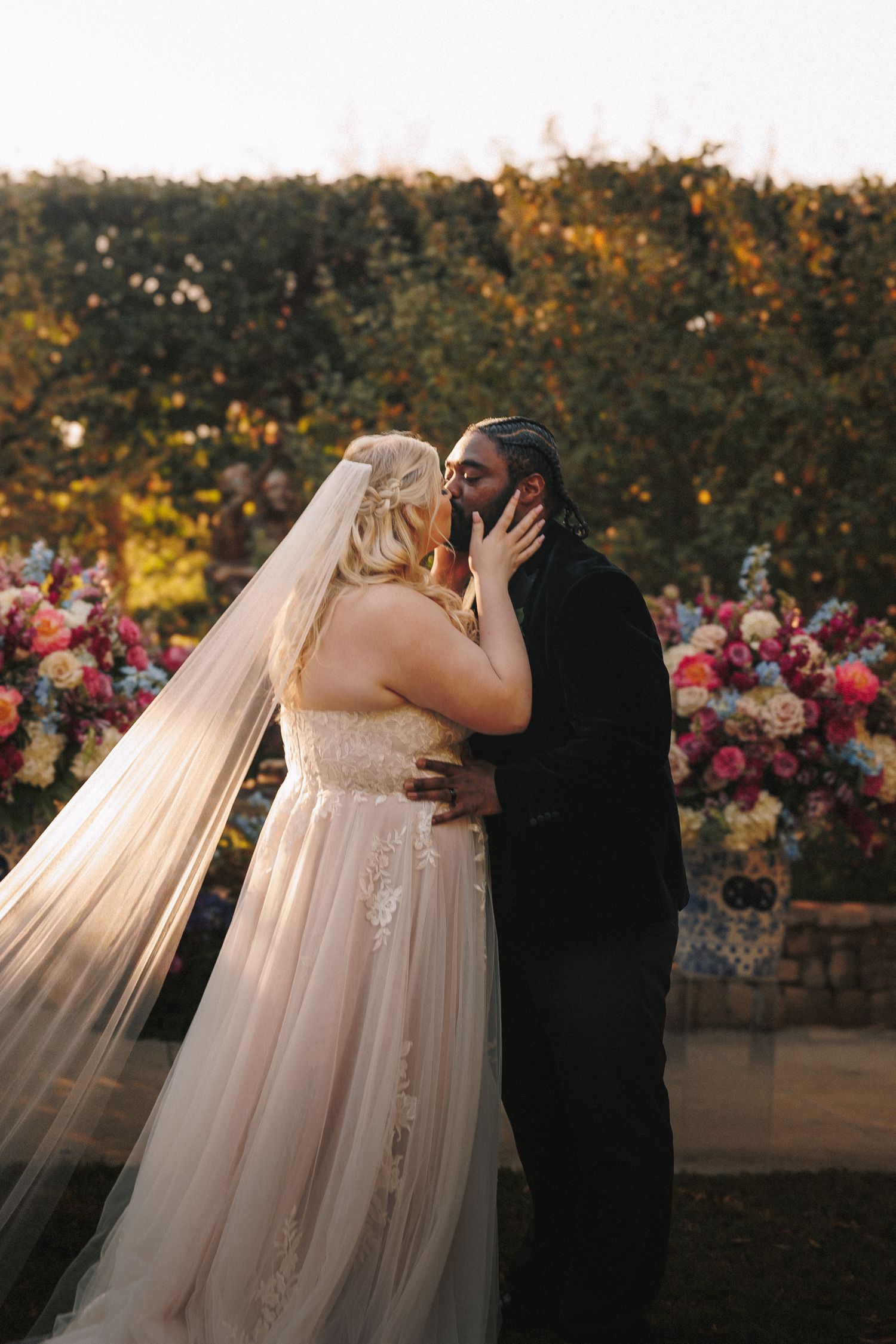 Bride and groom share first kiss surrounded by colorful floral arrangements at outdoor ceremony.